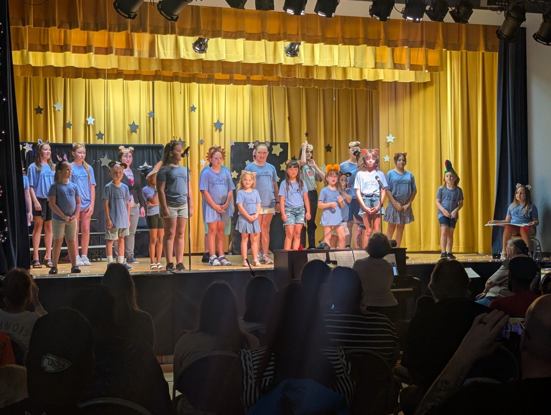 Children on a school stage under yellow curtains, singing and standing in rows before an audience.