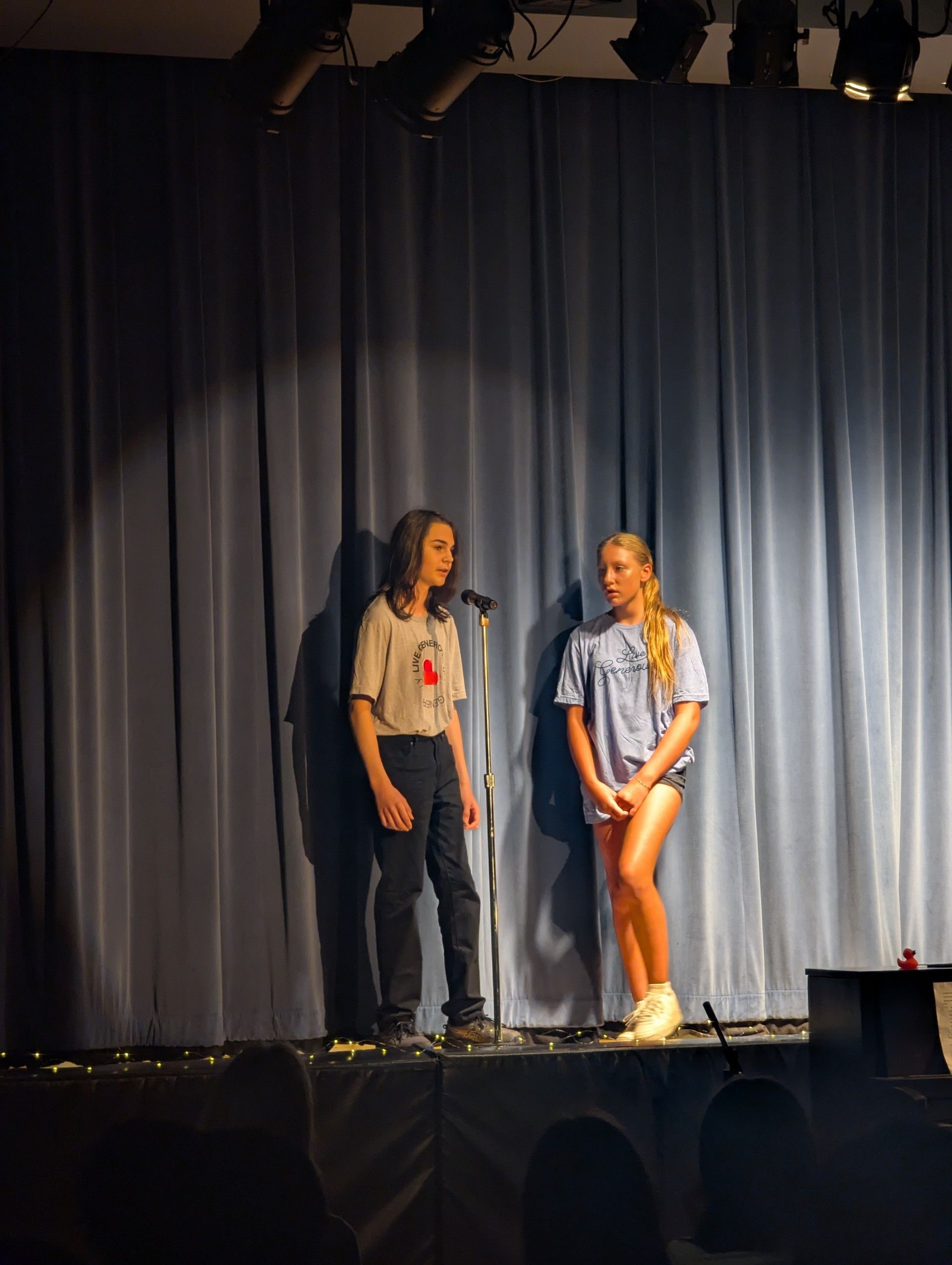 Two people on a stage in front of blue curtains, standing near microphones under stage lights.