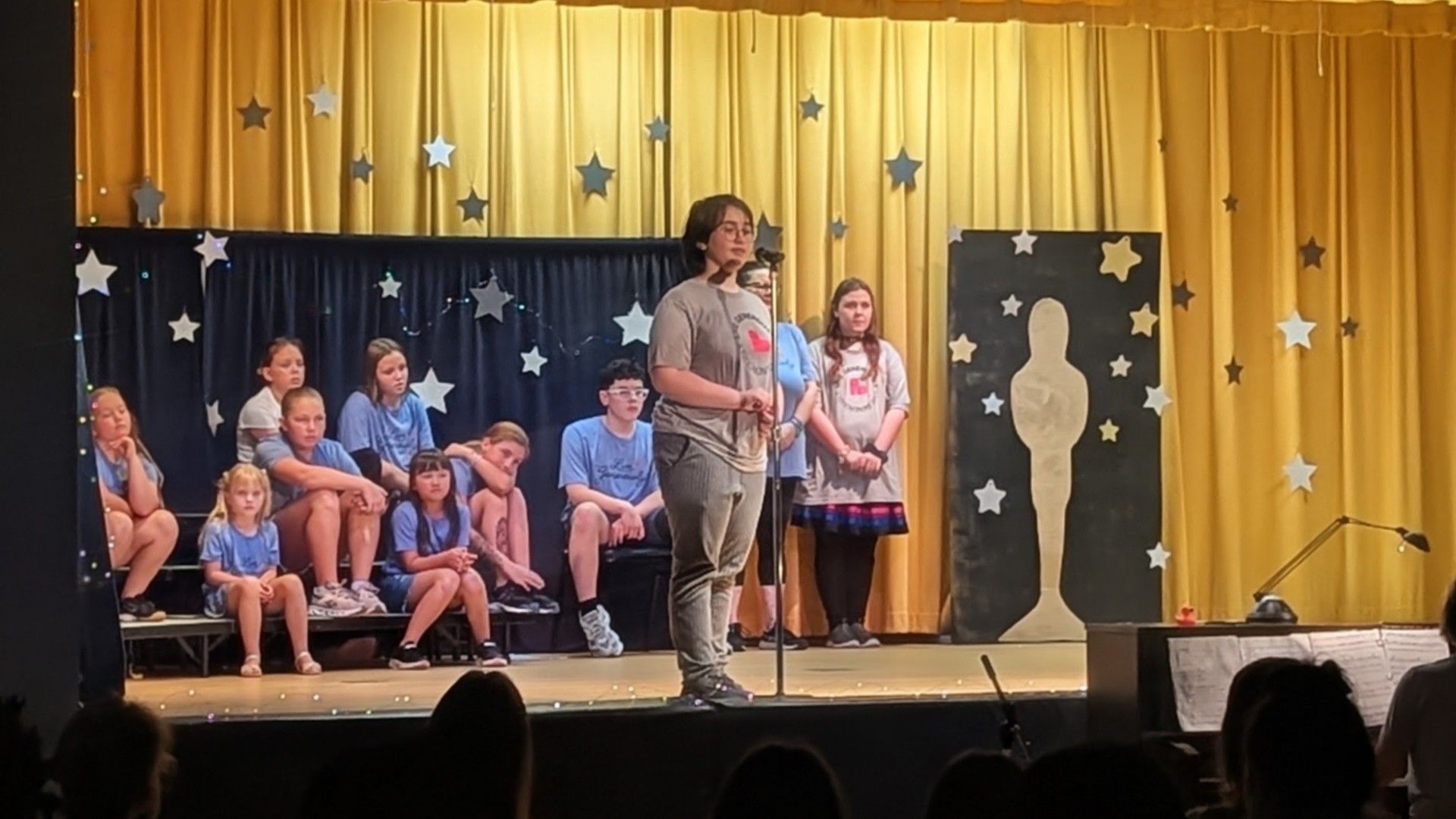 Children performing on a stage with gold curtains, stars, and a silhouetted figure backdrop