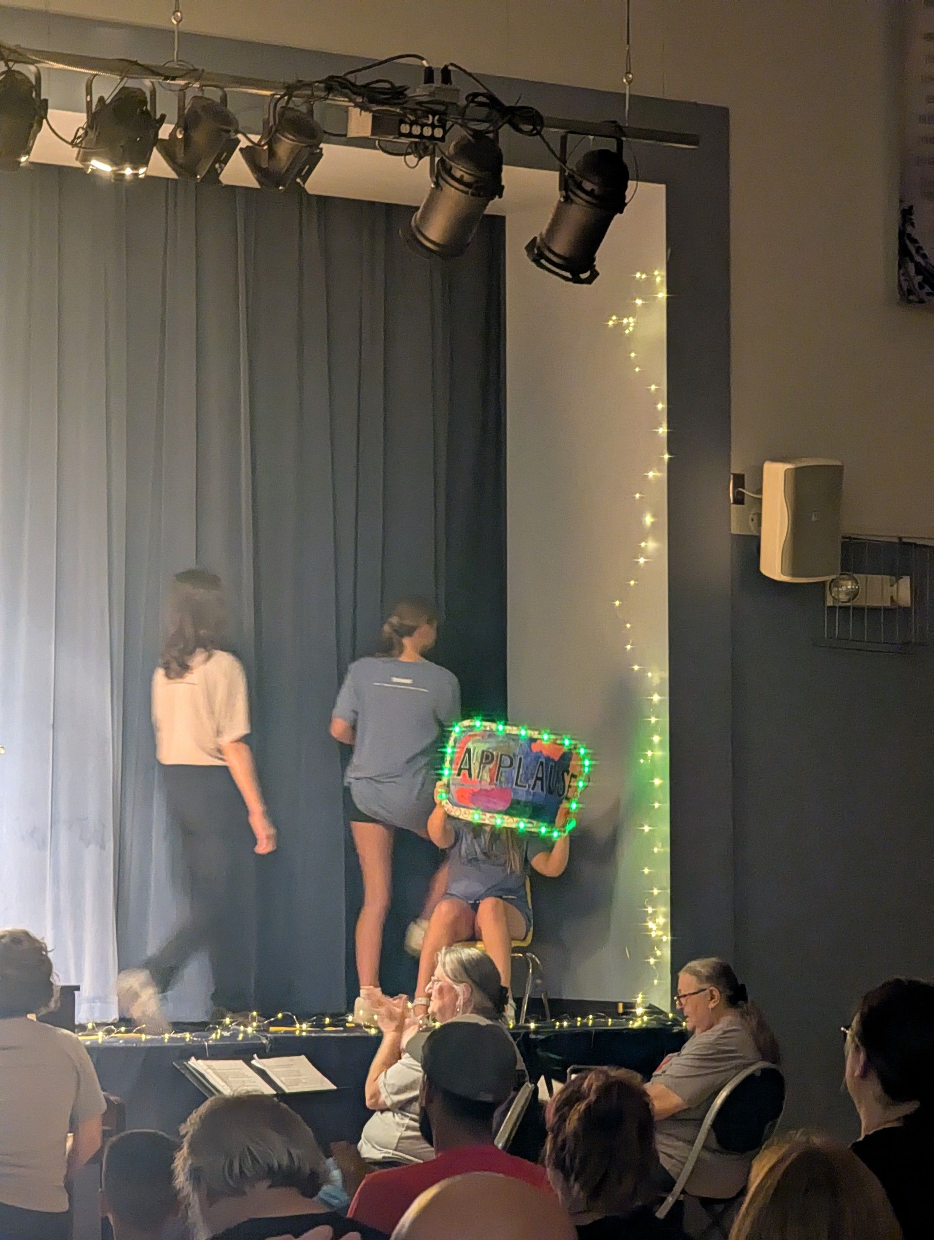 Two children onstage with a glowing toy; people watch from below beside a curtain and stage lights.