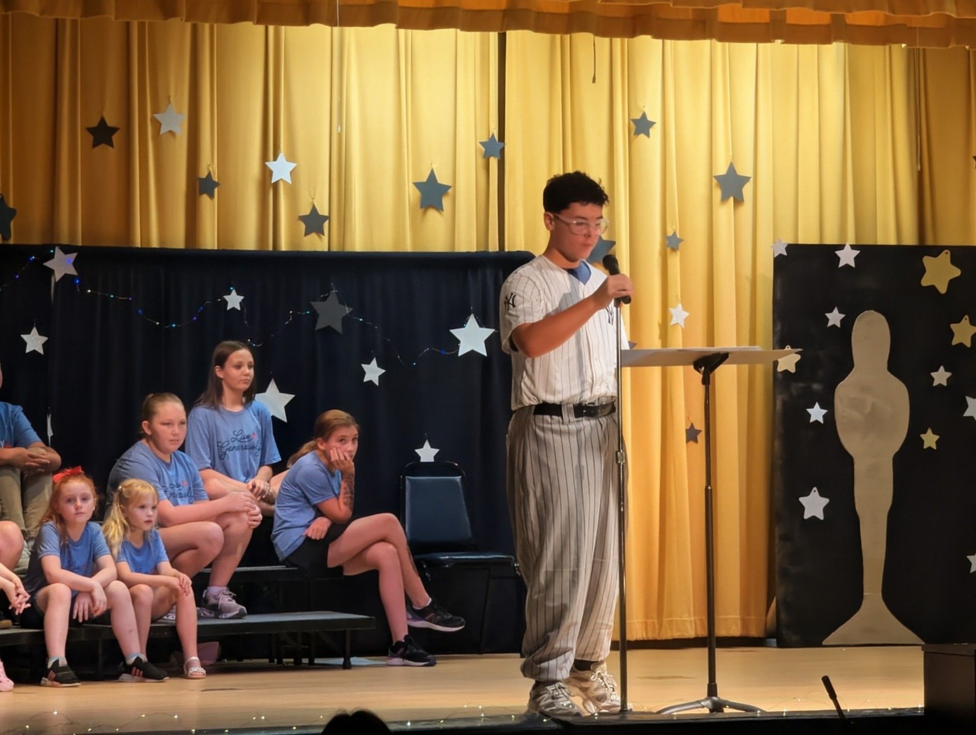 Man speaking at a podium on a stage with seated children and starry gold-and-black backdrop