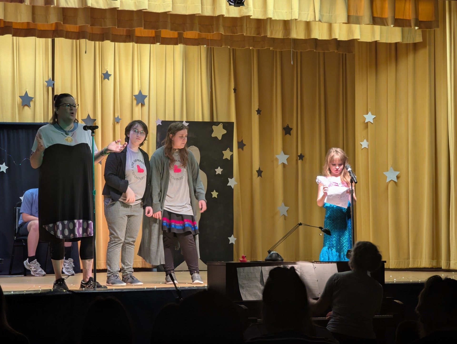 Children performing onstage in front of gold curtains, with an adult at left and audience silhouettes below.