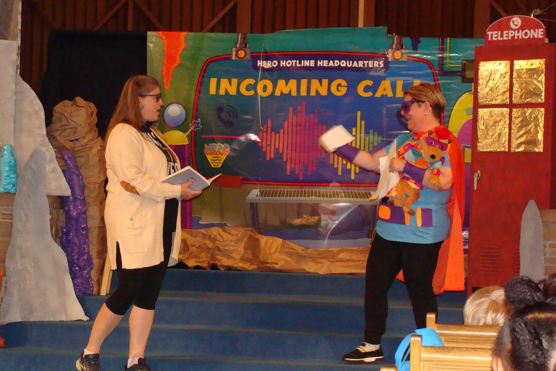 Two people on stage in front of a “Incoming Call” backdrop, one holding a microphone and cue cards.
