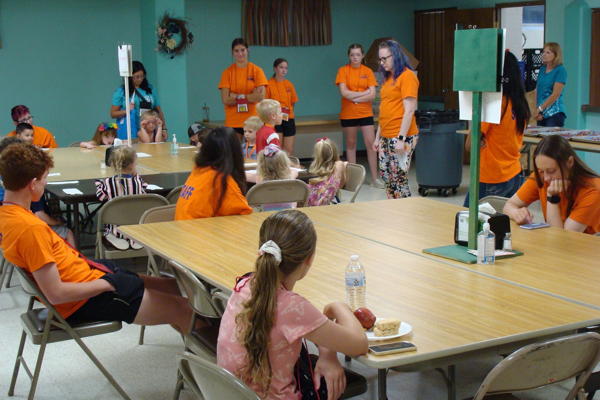 People in orange shirts stand and sit around a table in a community room, with a whiteboard and decorations.