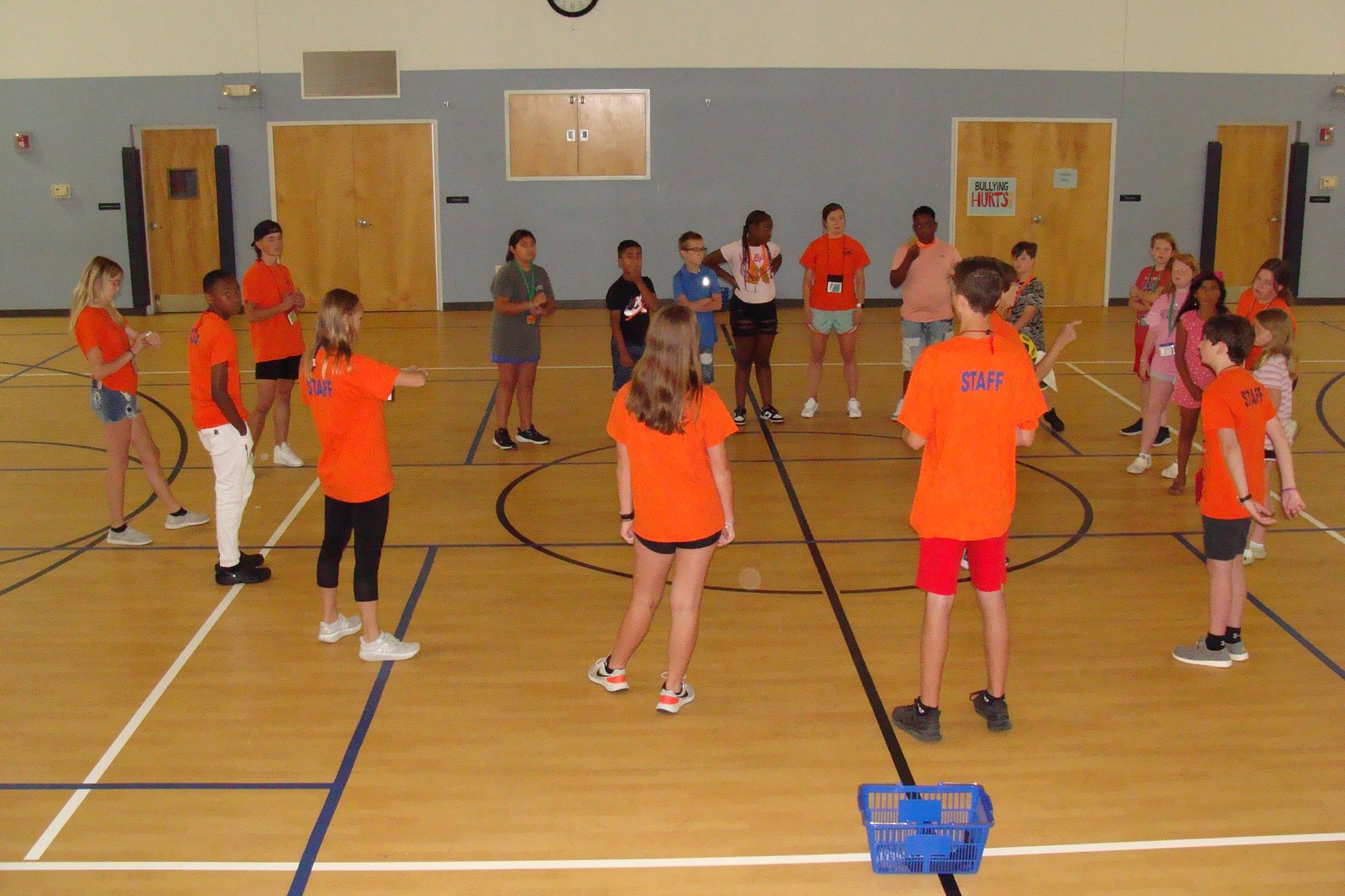 Children in orange shirts practice in a gym with floor markings and hockey sticks.
