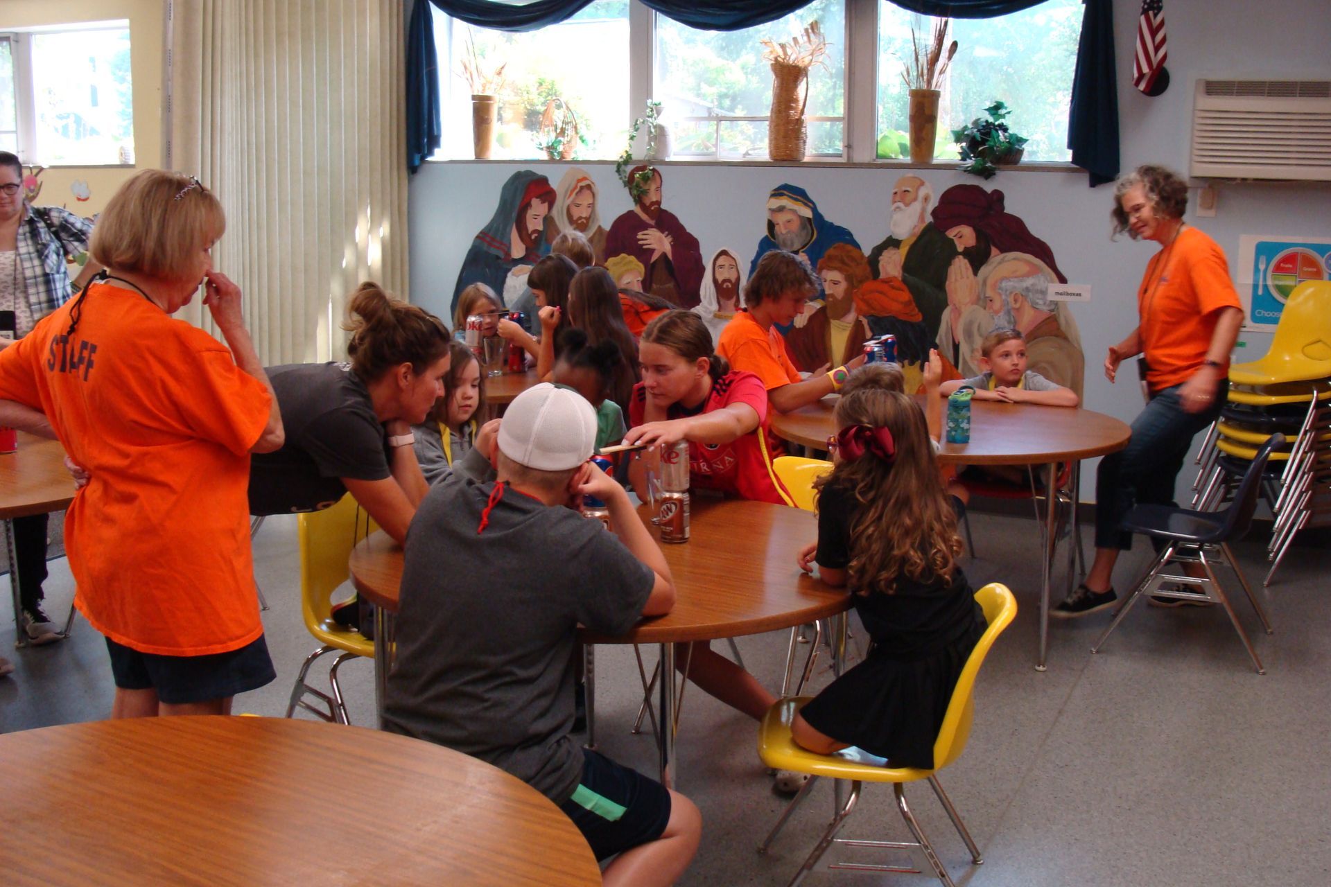 Children and adults seated around tables in a bright room, doing an activity with colorful artwork displayed behind them