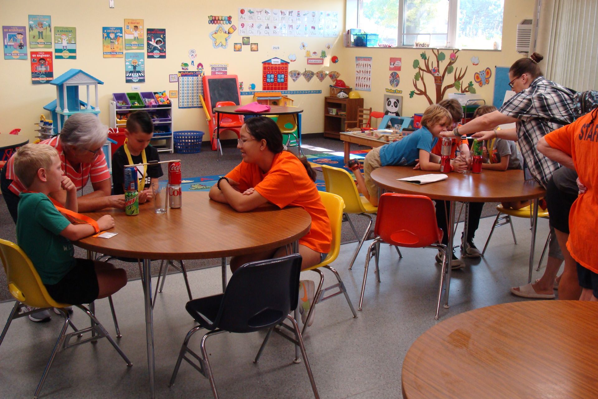Children sit at tables in a colorful classroom, working and talking during a group activity.