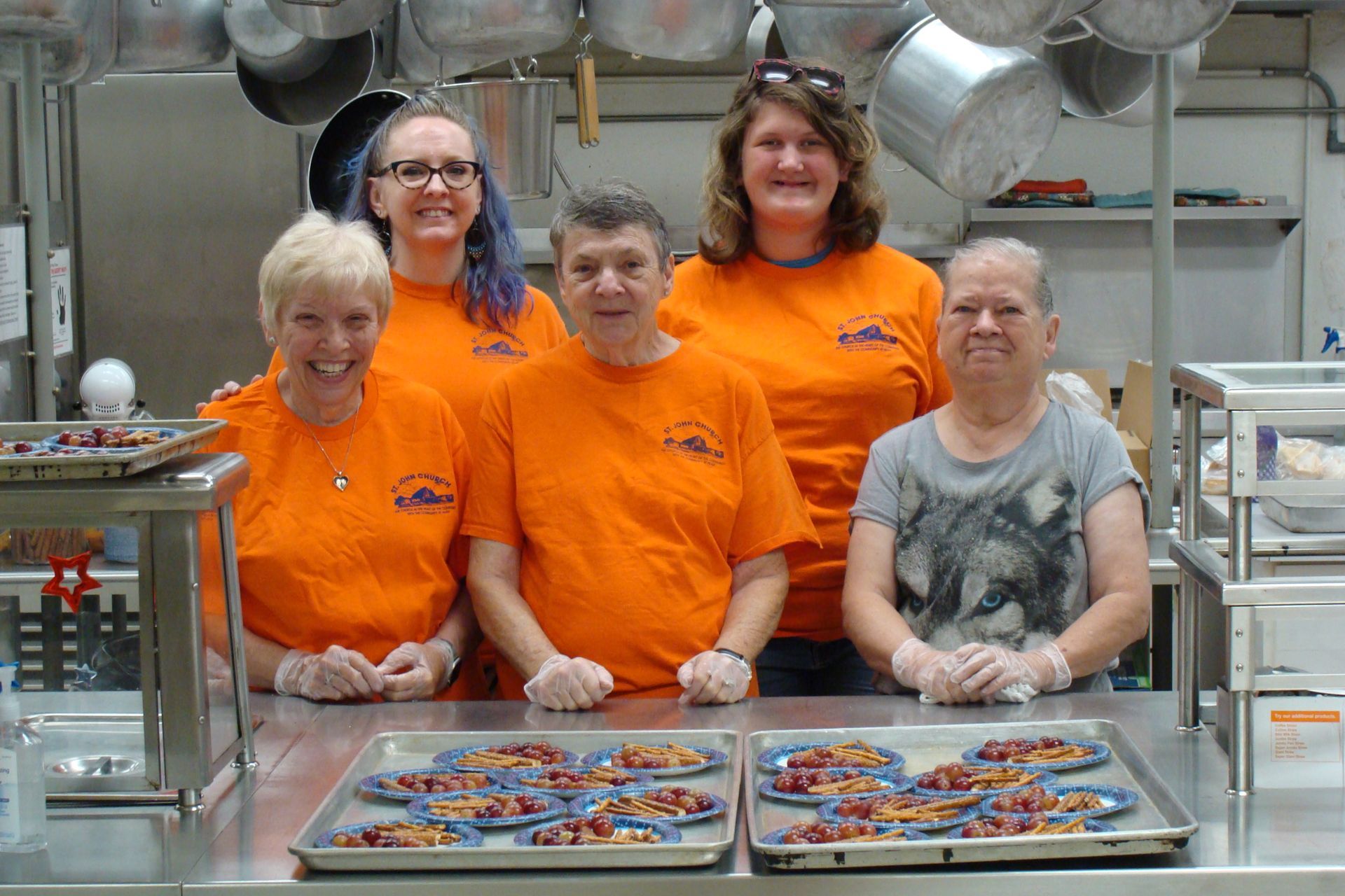 Five workers in orange shirts and gloves stand behind trays of colorful food in a factory kitchen.