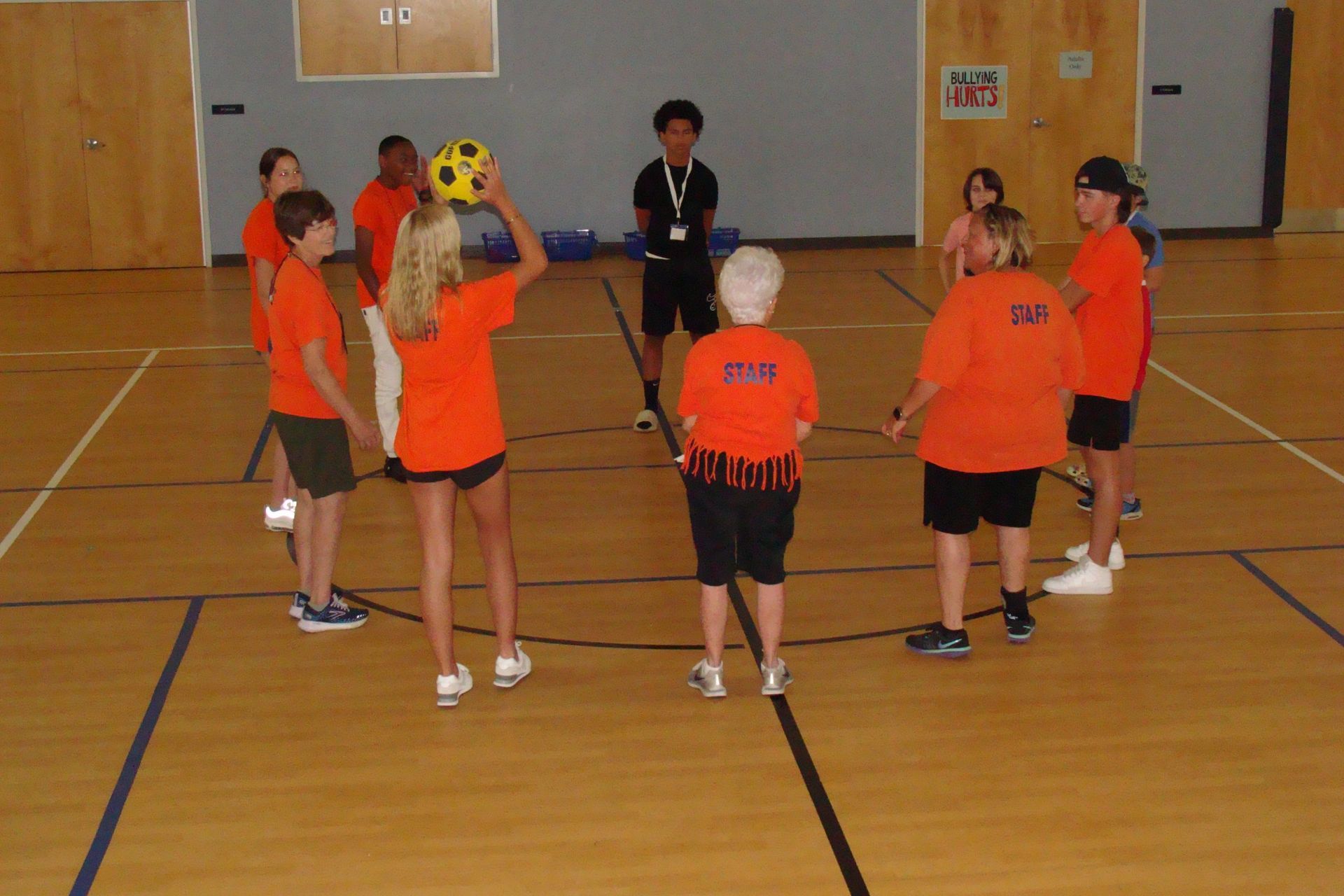 Children in orange shirts stand in a gymnasium, gathered in a circle around a coach.