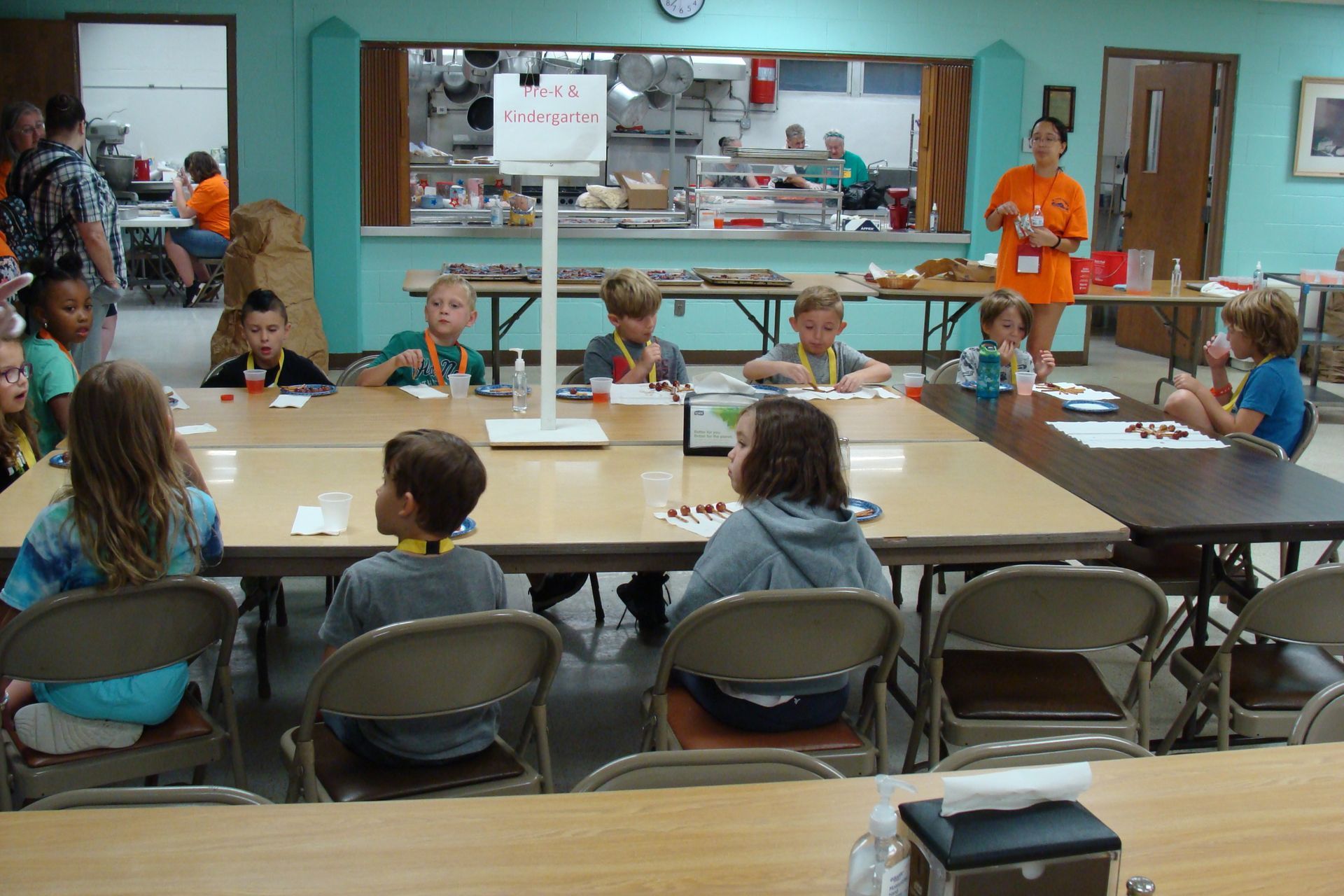 Children seated around tables in a classroom, with a teacher near a front counter and kitchen area.