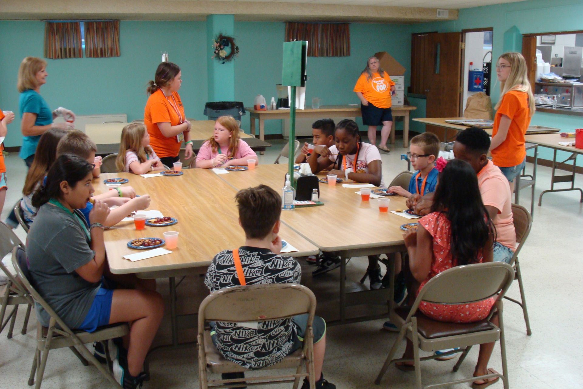 People sitting around a table in a community room, with two adults in orange shirts nearby.
