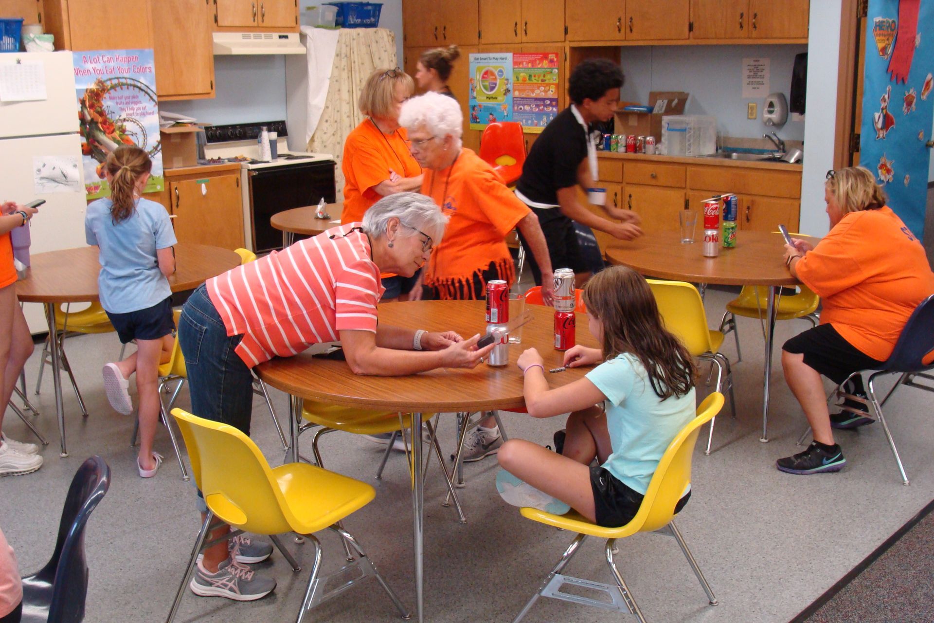 People around a table in a classroom kitchen, preparing food with orange and yellow chairs.