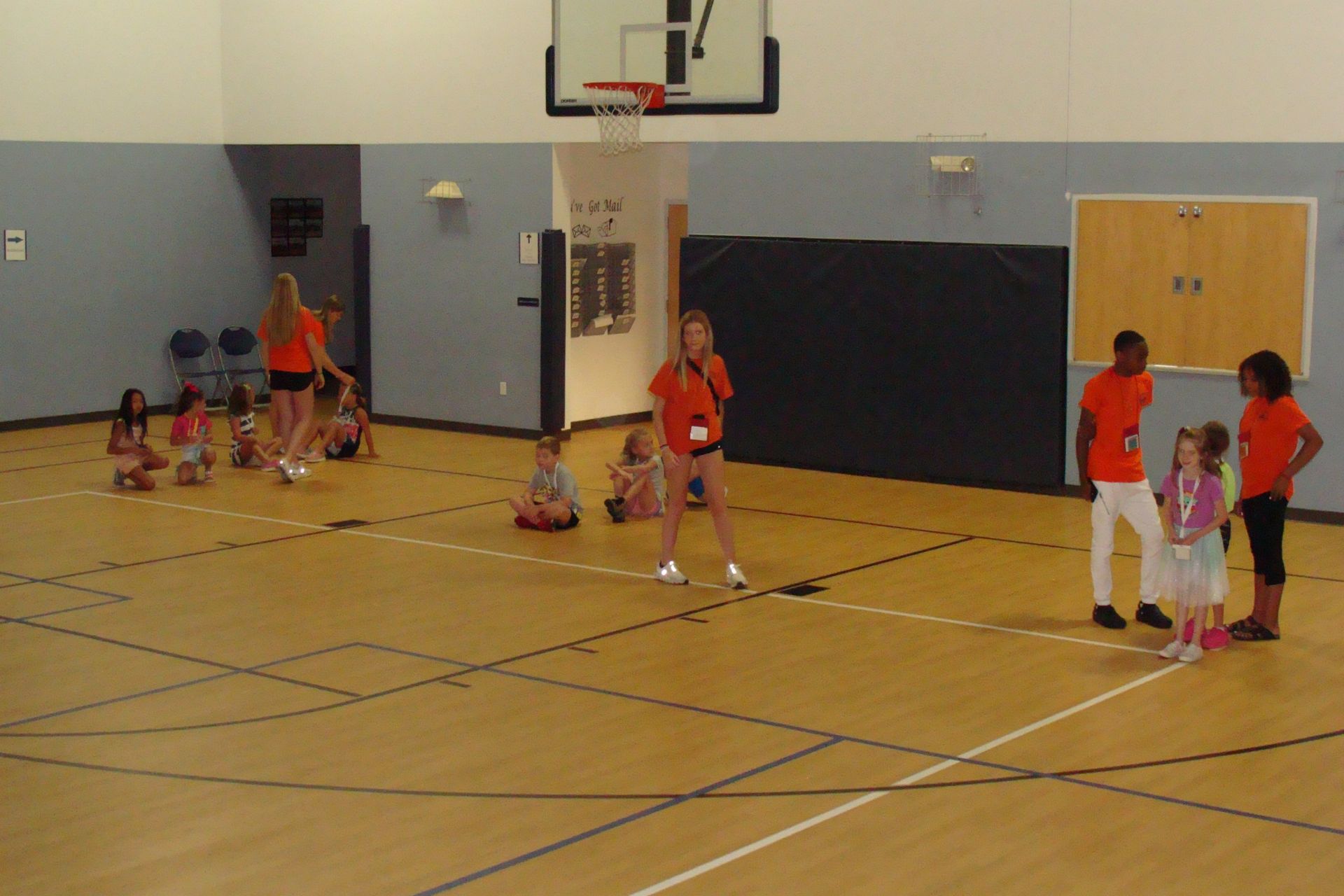 Children in a gym playing around a basketball hoop, some in orange shirts.