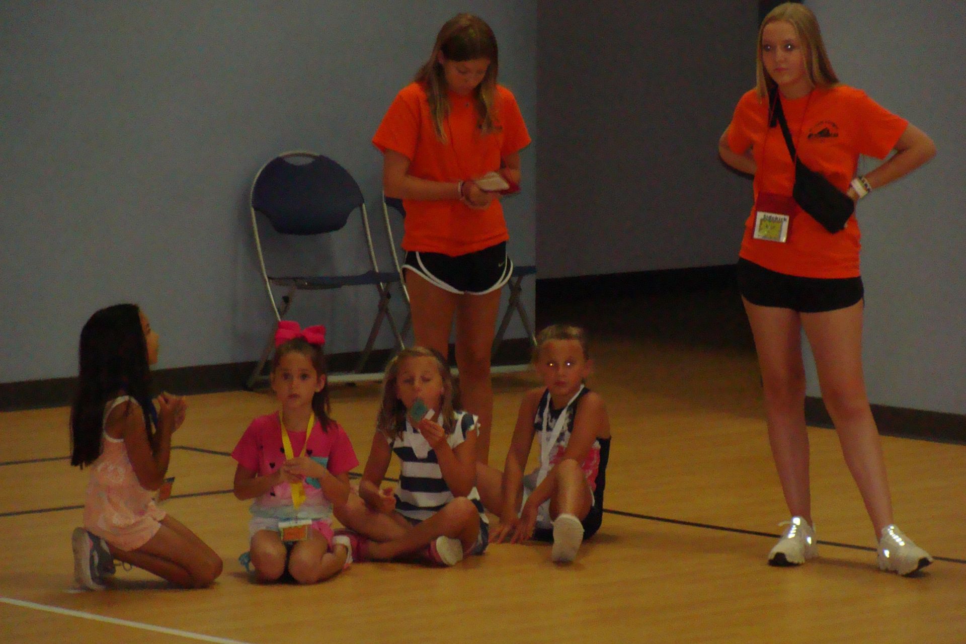 Children seated on a gym floor with two orange-shirted coaches nearby, one standing and one bending over.