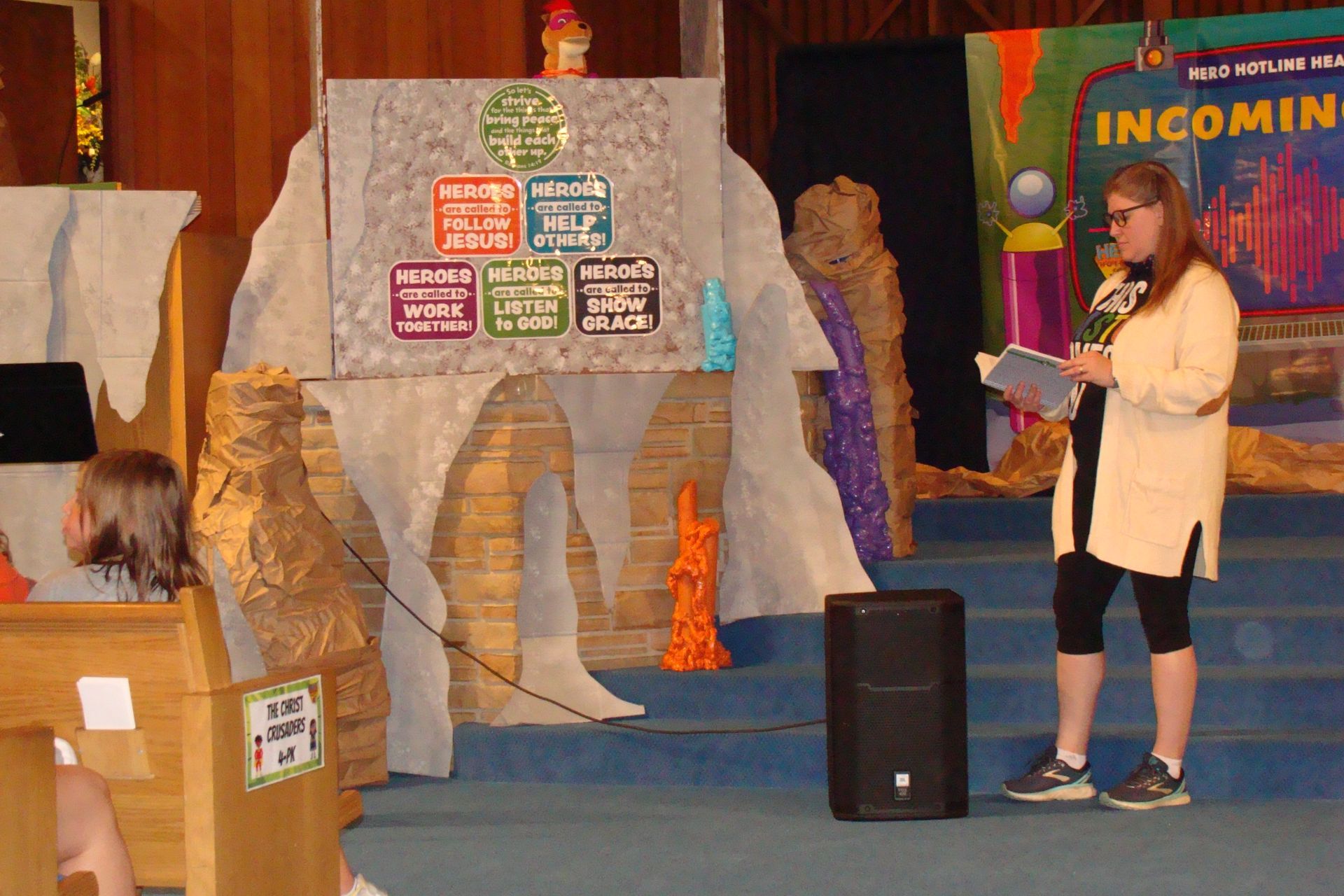 Woman speaking at a decorated stage beside a rock-like display and “Incoming” sign
