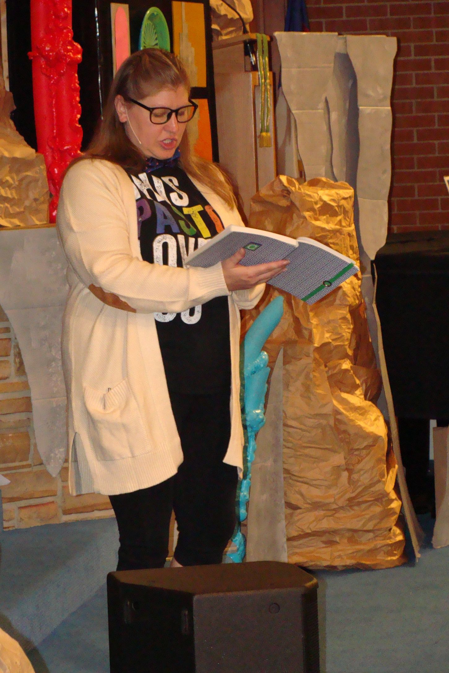 Woman reading from a blue booklet beside a gold and gray display on a stage