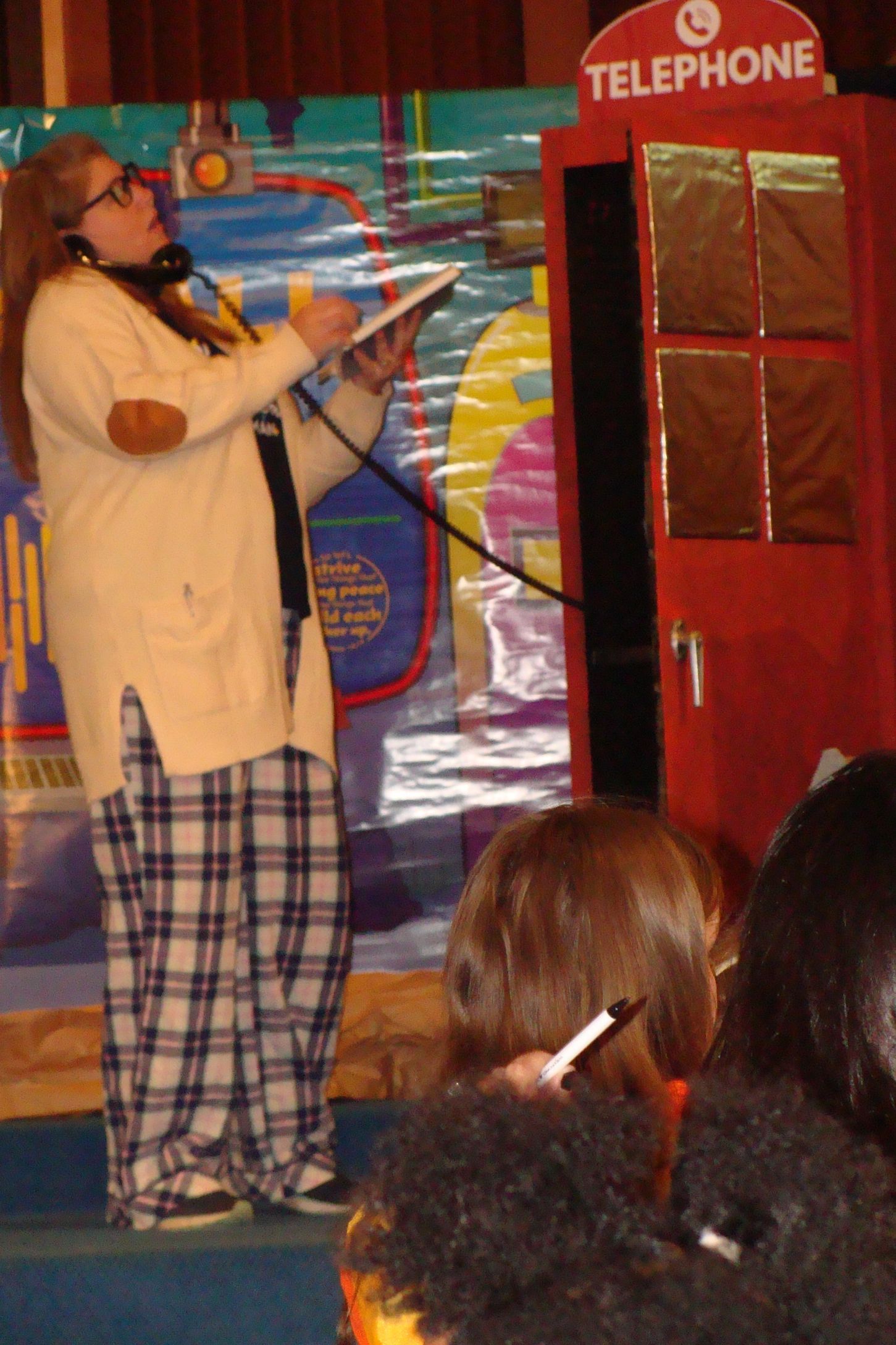 Woman speaking into a red telephone booth on a stage, holding papers while an audience watches