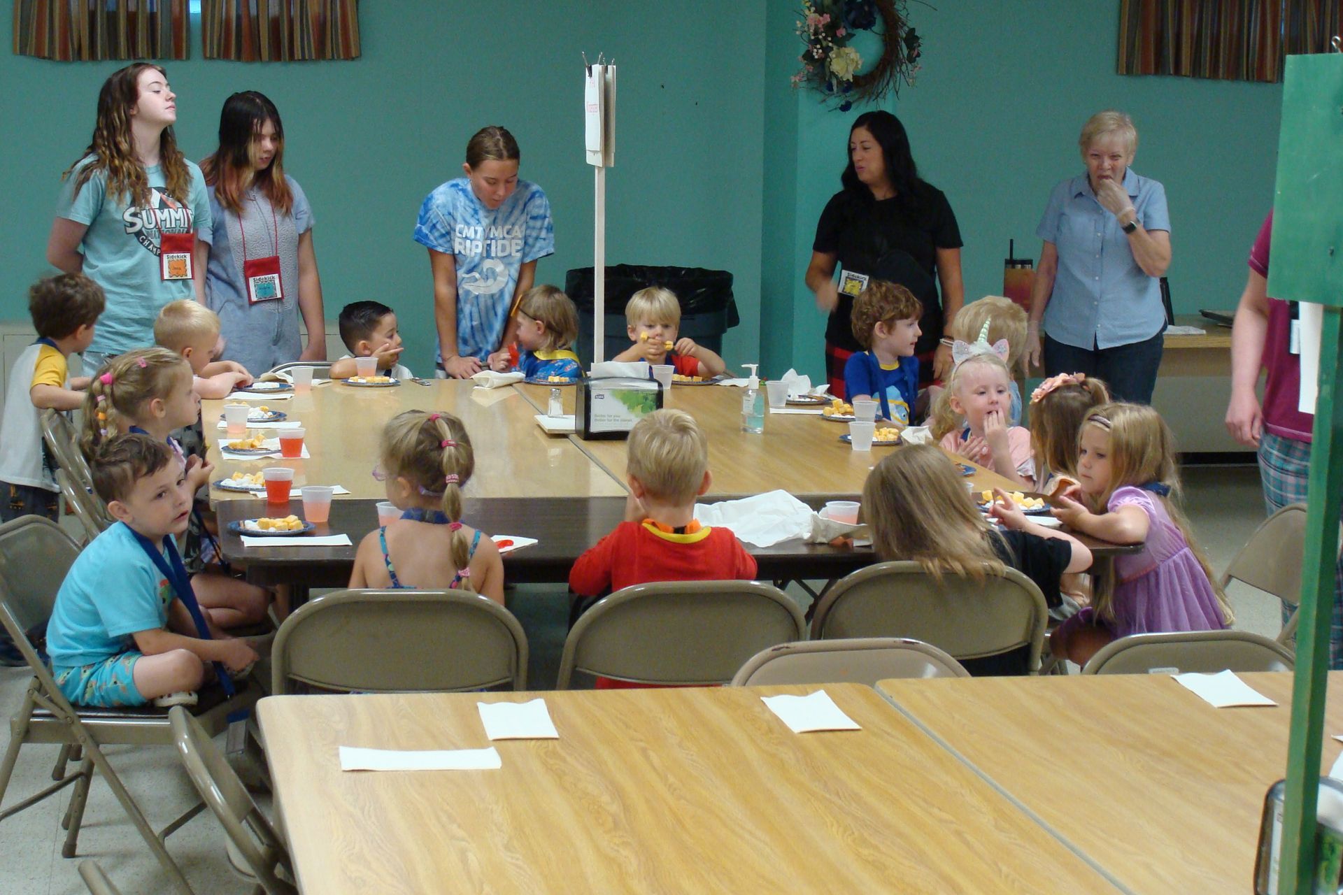 Children seated at tables in a classroom, with adults standing nearby during a group activity.