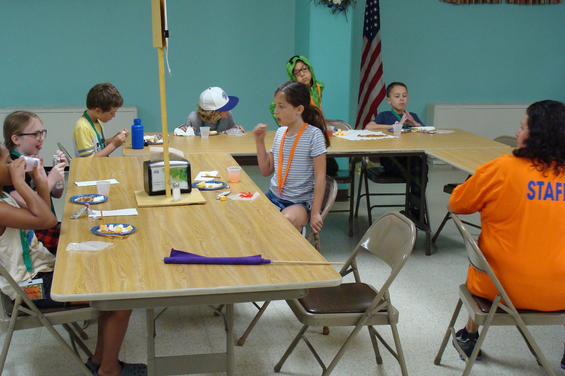 People seated at tables in a community room, with a staff member in an orange shirt nearby.
