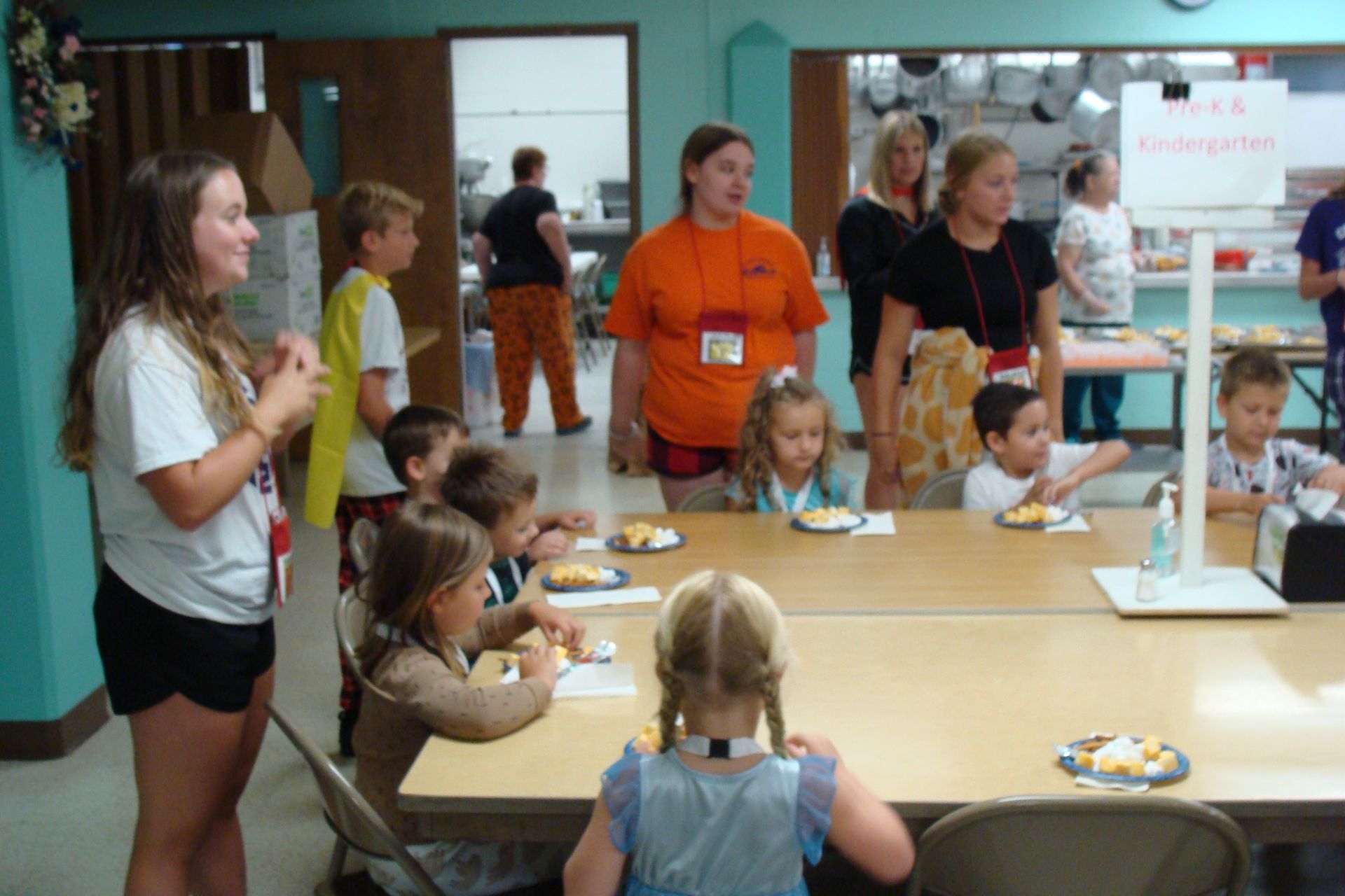 Children and adults gathered around a table in a brightly colored school or community room.