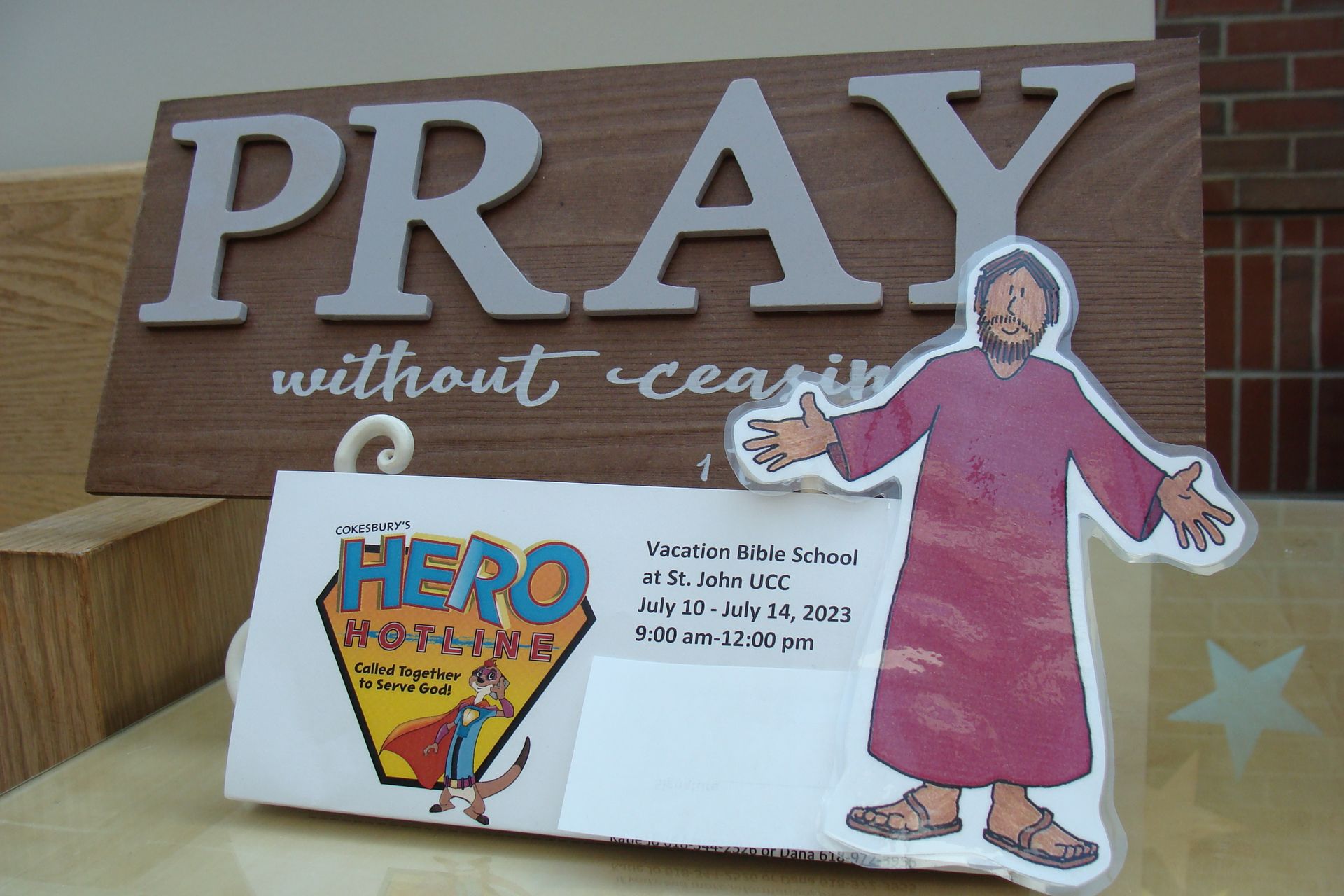Wooden sign reading “PRAY without ceasing” beside a cutout of a girl in a pink dress and a “HERO” notice card