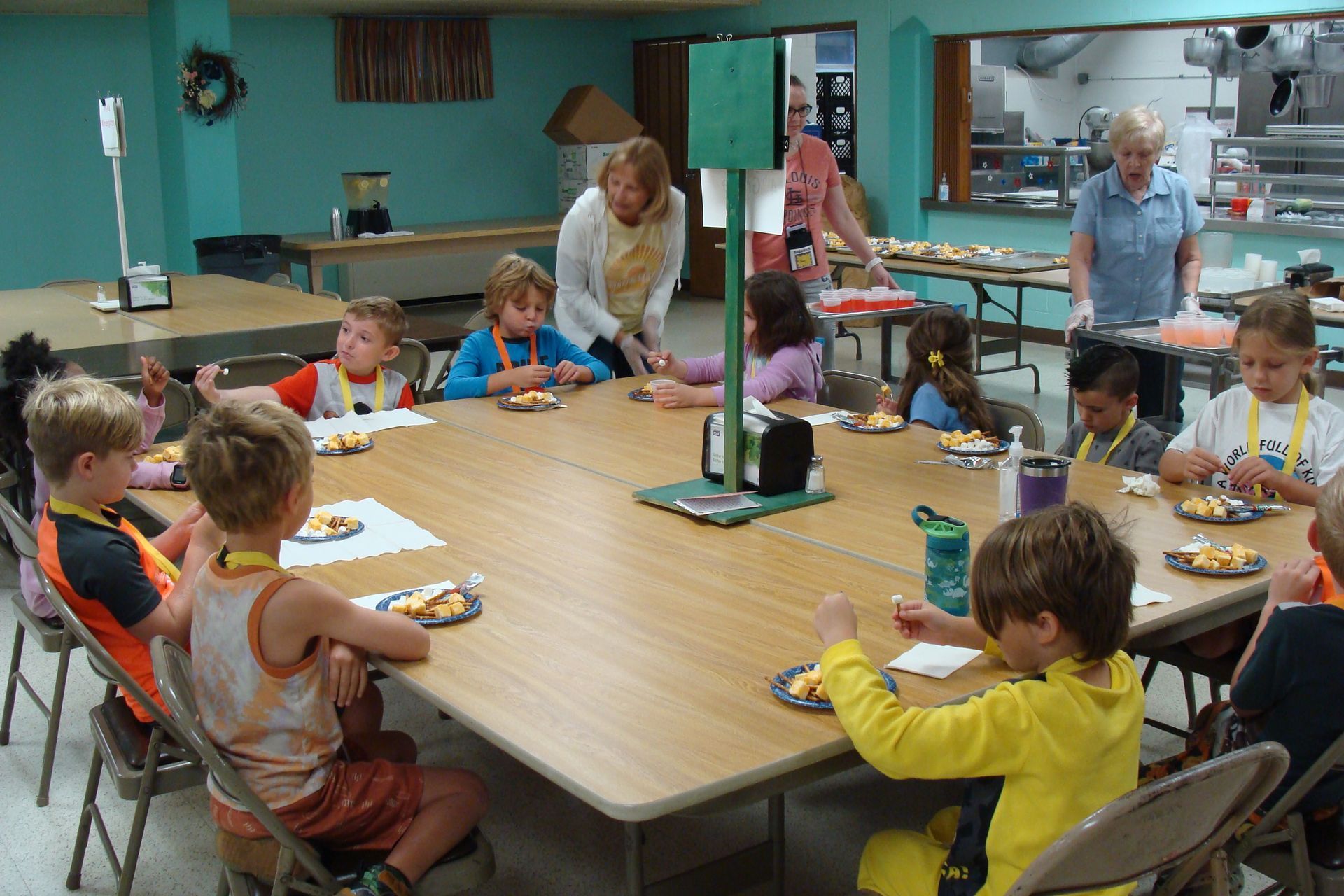 Children eating at a large round table in a bright cafeteria with adults serving food.