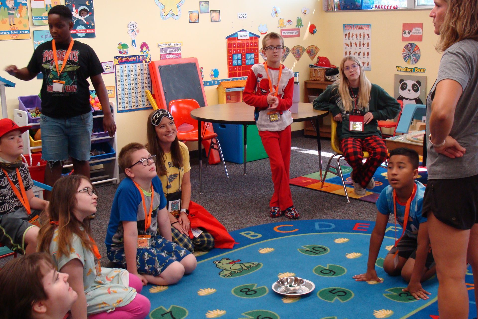 Children sit in a colorful classroom while a teacher demonstrates an activity at the front.