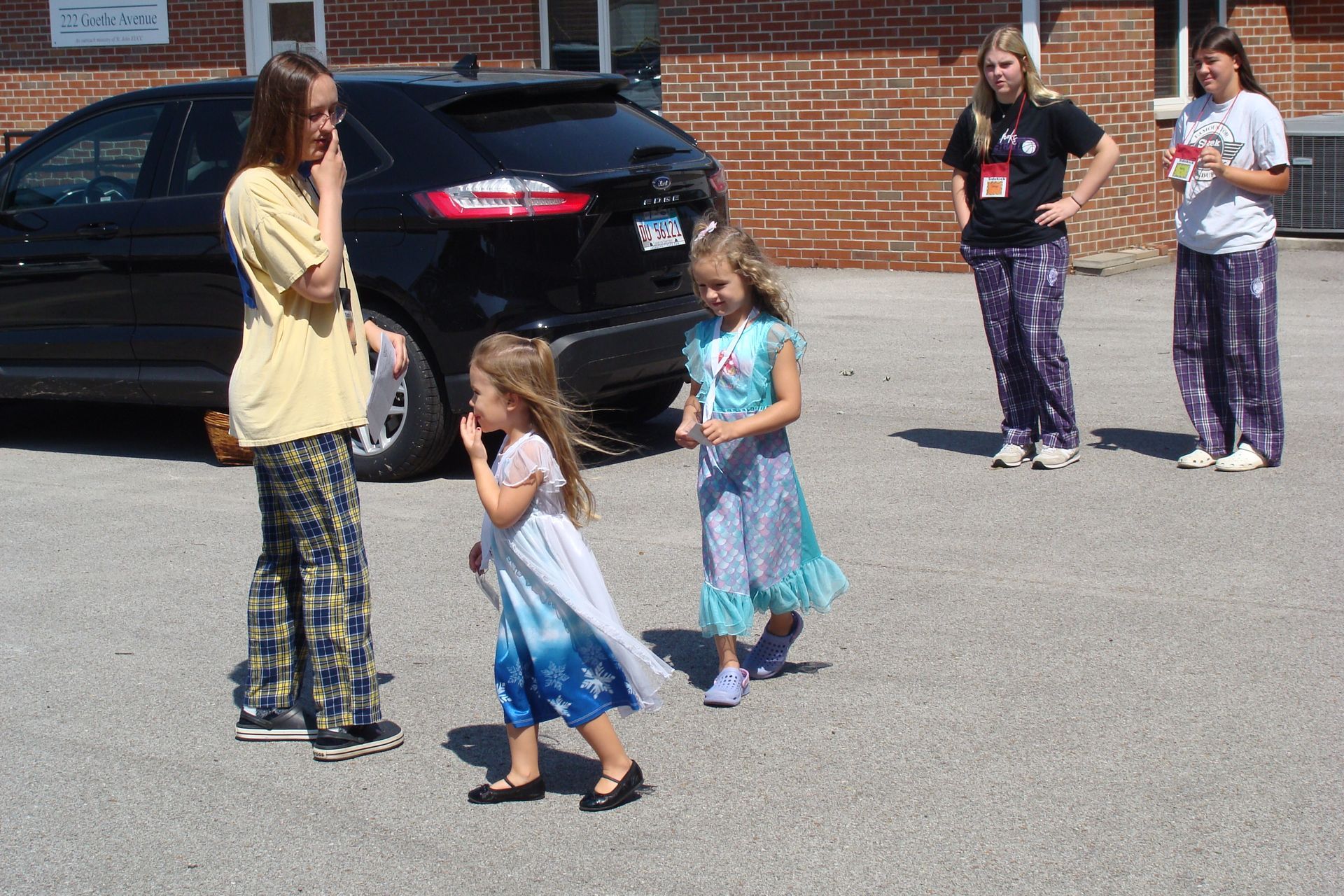 People standing in a parking lot near a black car, with two children and two adults chatting.