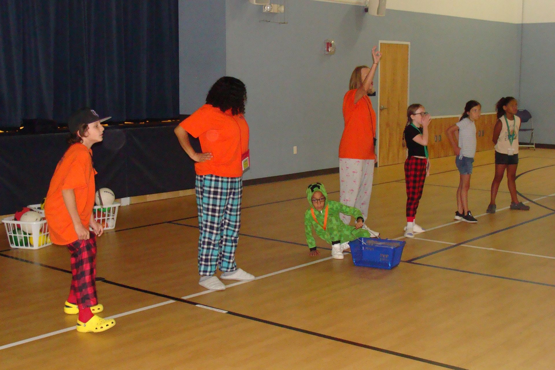 Children in orange shirts standing in a gym during a school activity, with one child in green sitting on the floor.