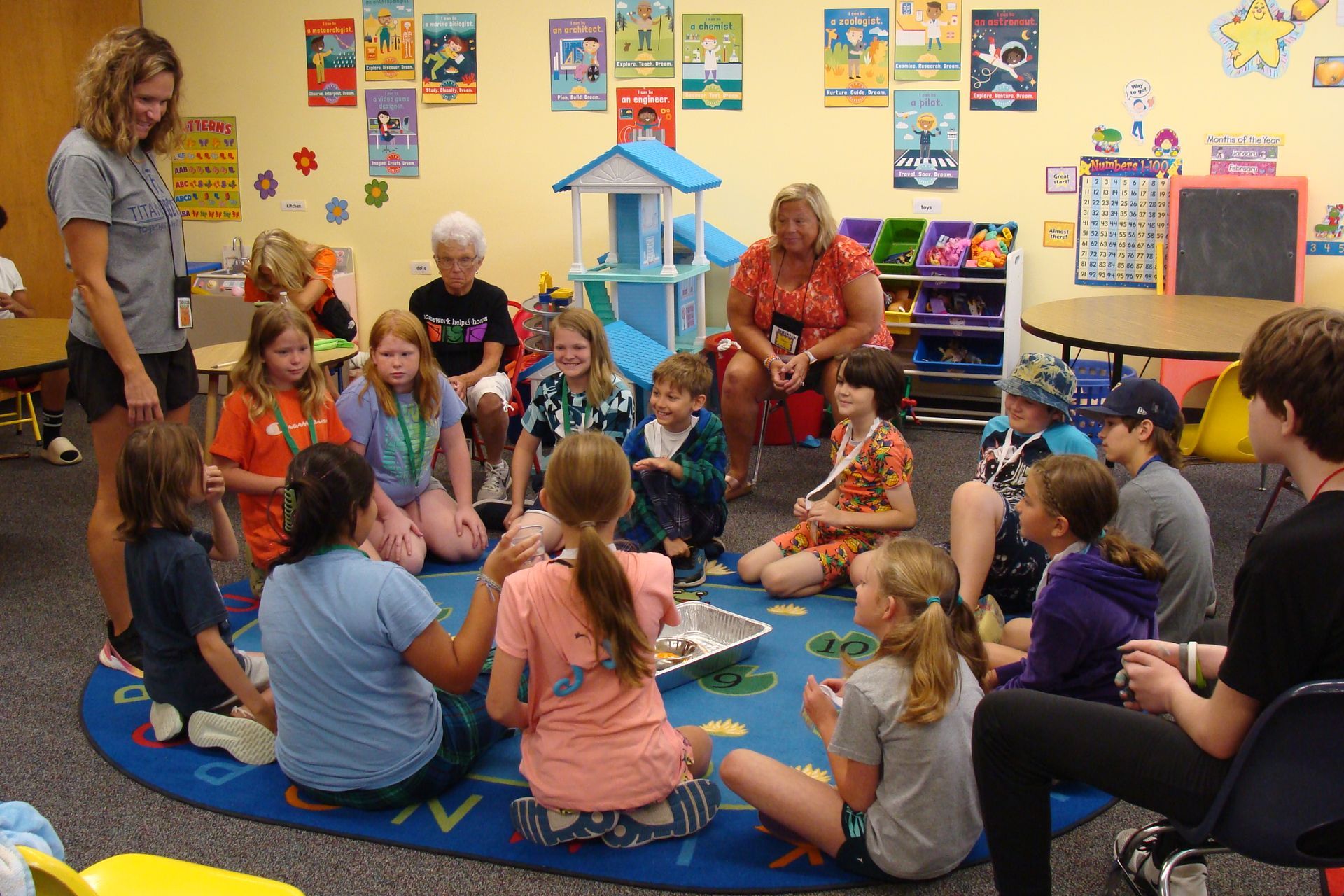 Children sit in a circle on a classroom rug, listening to a story with teachers nearby.