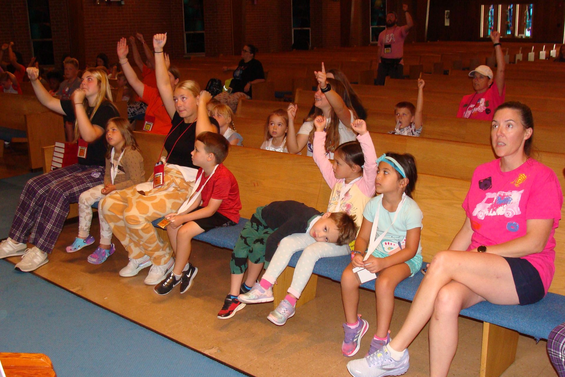 Children sitting on chairs in a hall, raising hands during a group activity