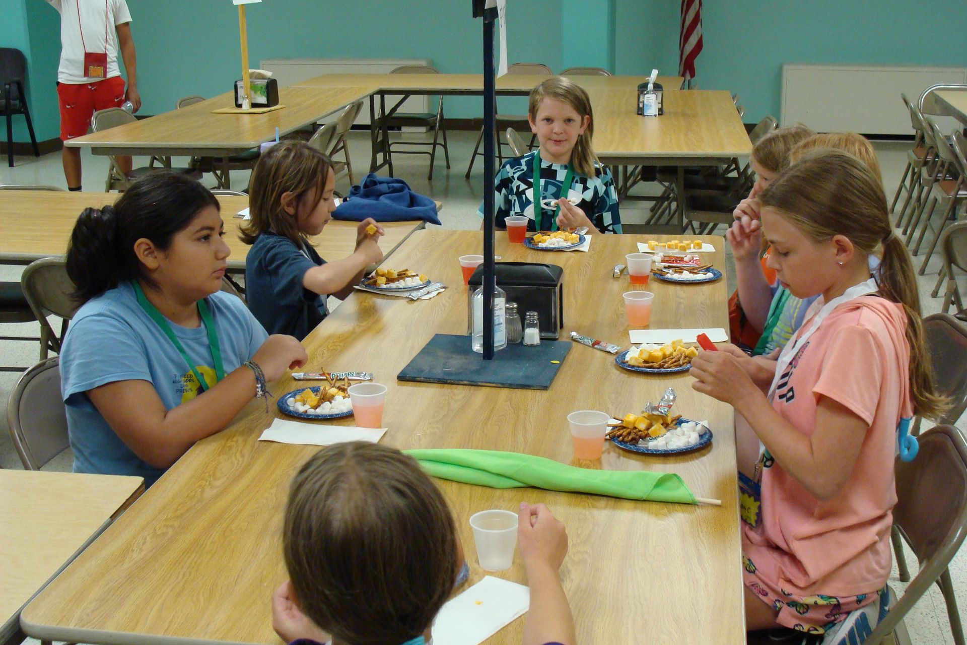 Children seated around a cafeteria table sharing a snack, with plates and drinks in a school gym or lunchroom.