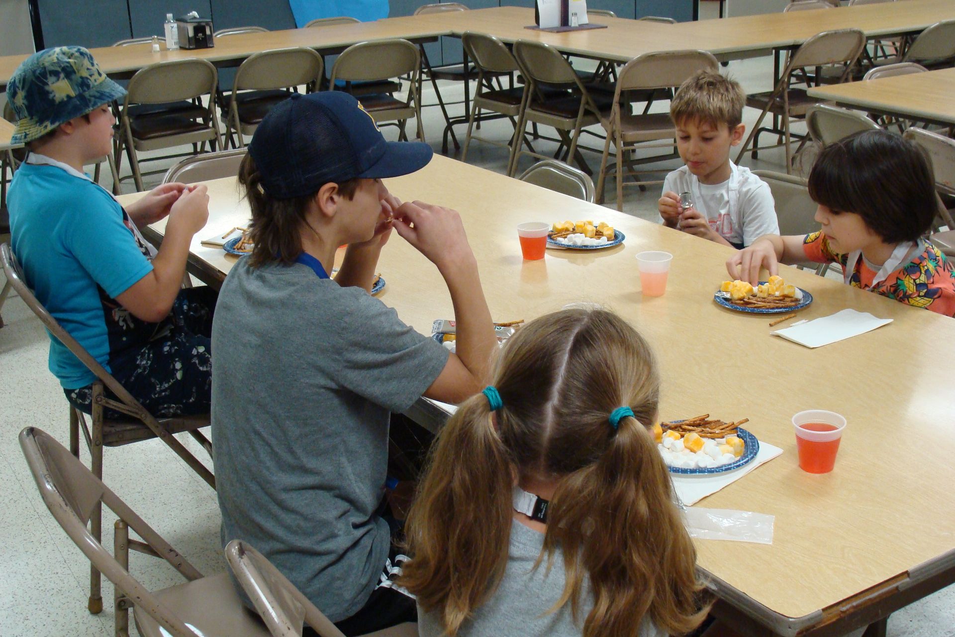 Children seated around a cafeteria table eating lunch and talking in a classroom.