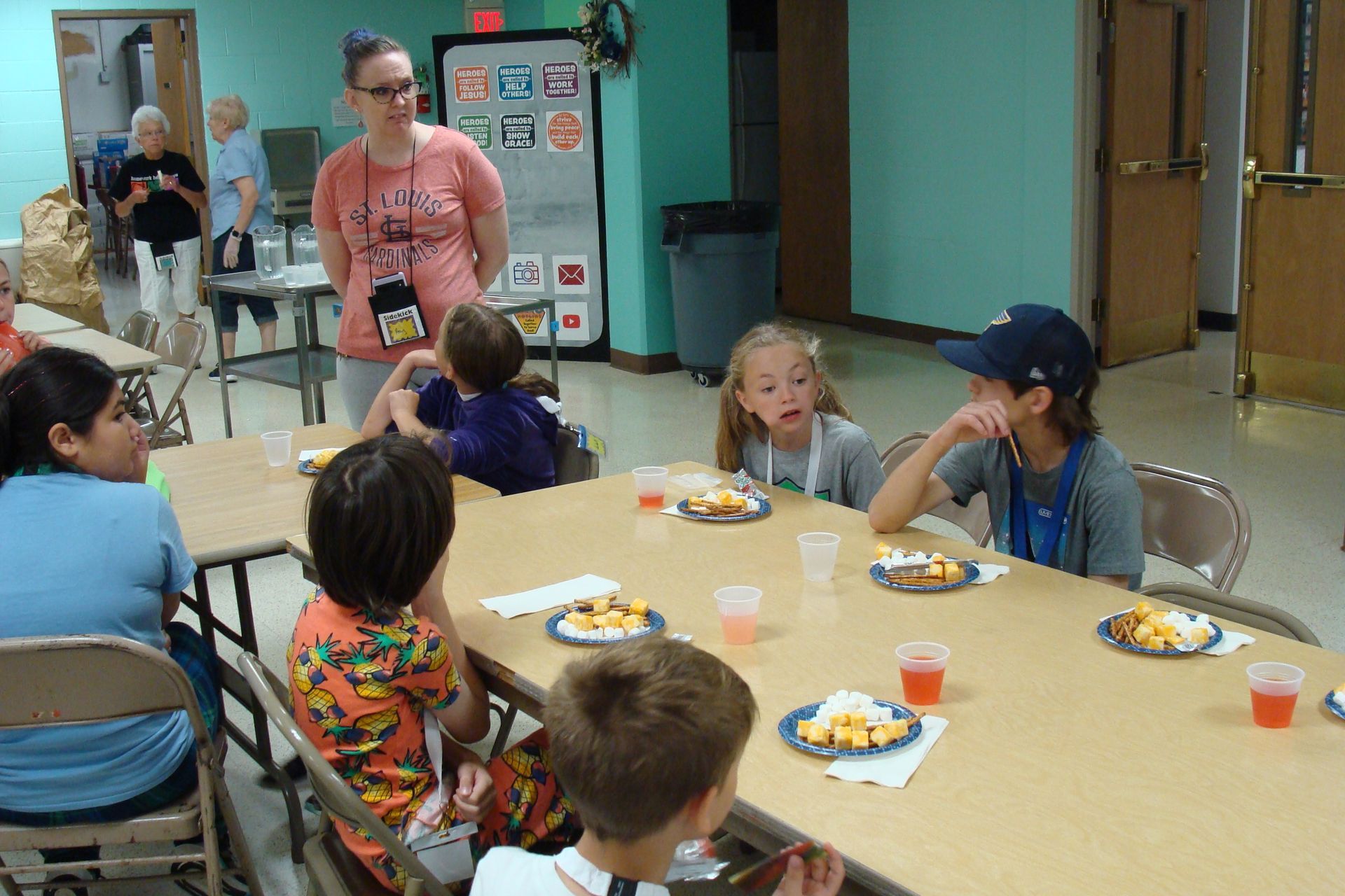 Children seated around a cafeteria table eating snacks while a woman stands nearby in a school hallway.