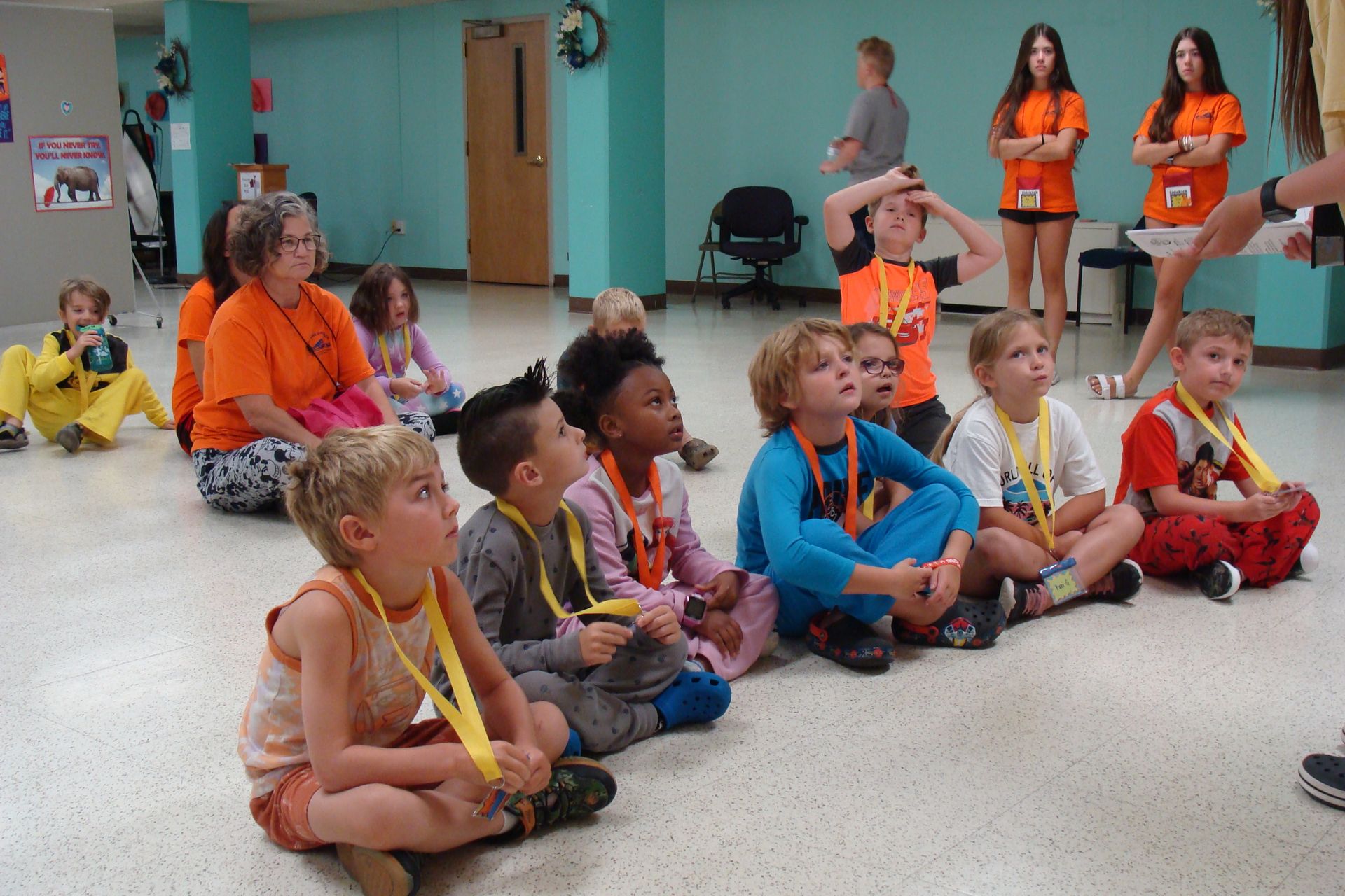 Children sitting cross-legged in a classroom watching a seated speaker, with volunteers standing nearby