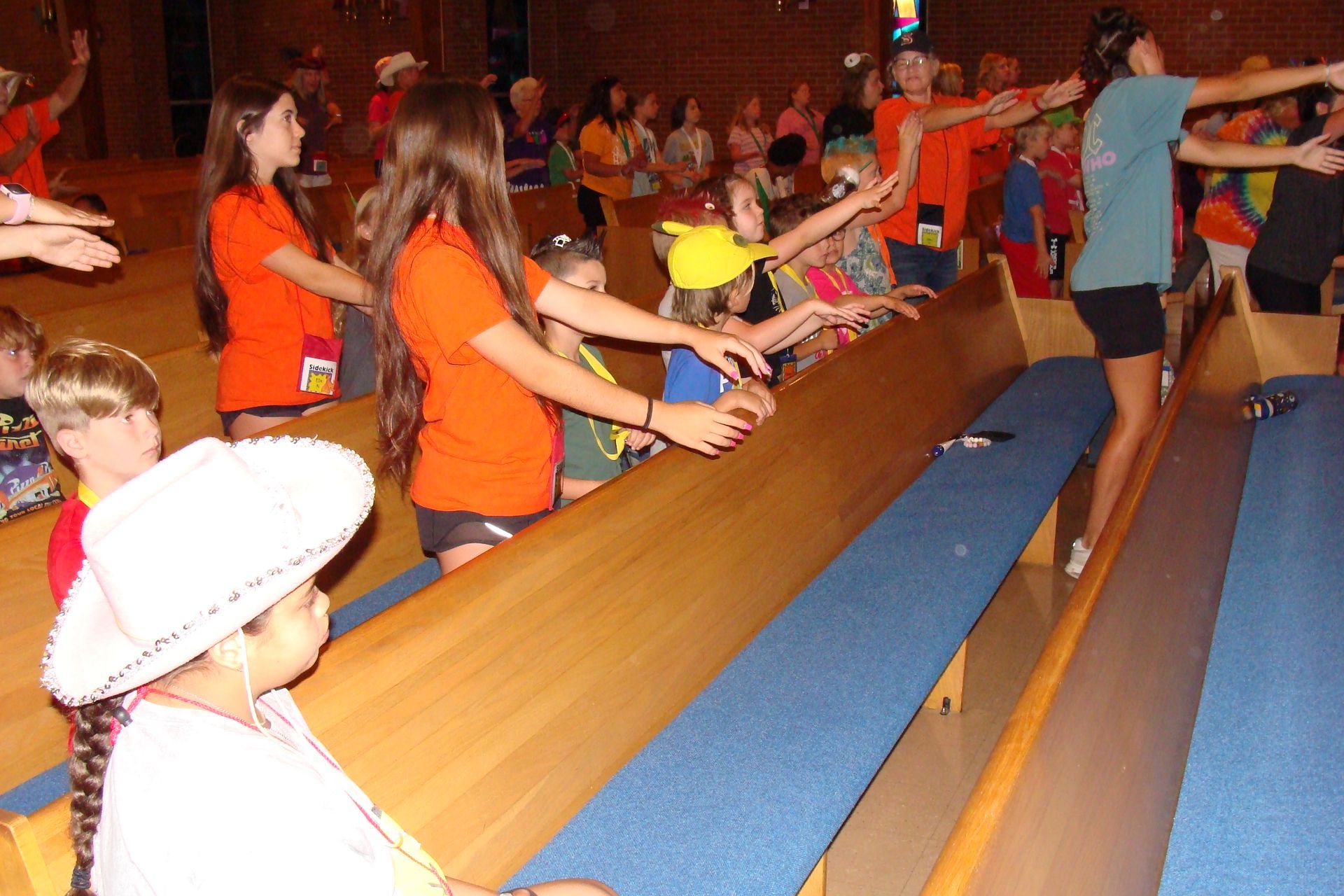 Children in orange shirts stand by long wooden benches in a busy indoor hall