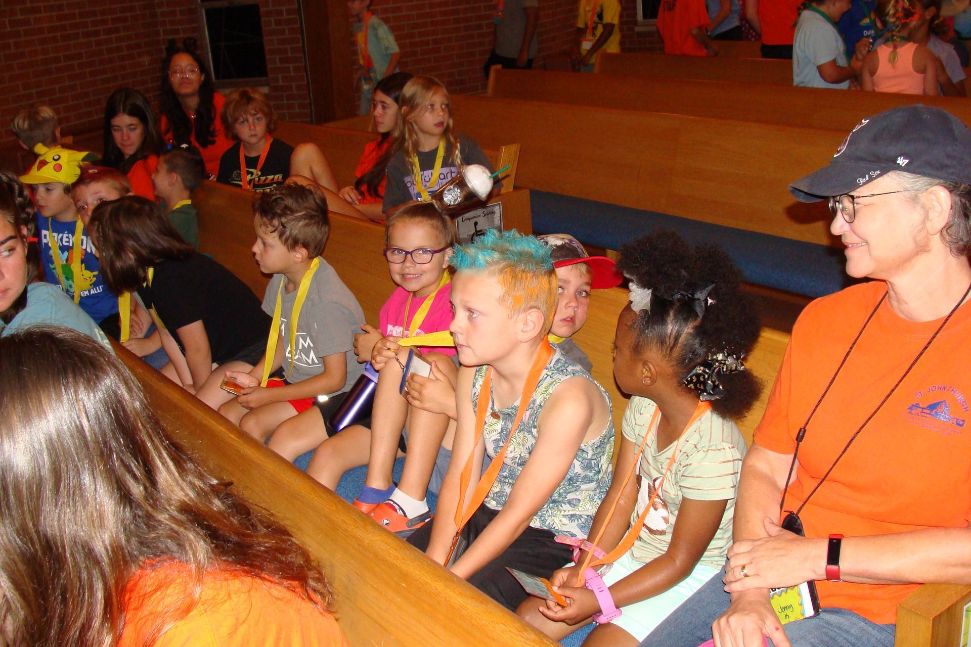Children seated in rows at an indoor event, listening attentively under warm lighting