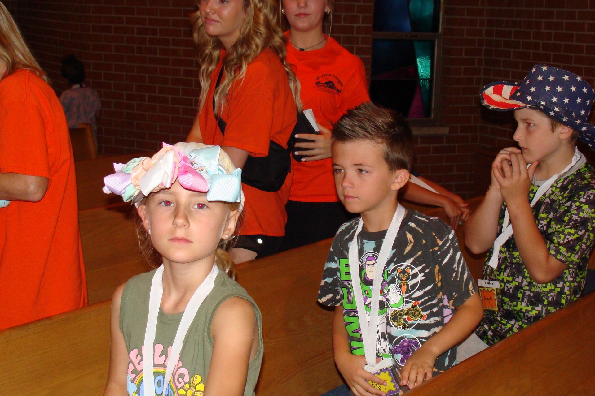 Children wearing leis sit at a table in a dim indoor room, one with a floral headband.