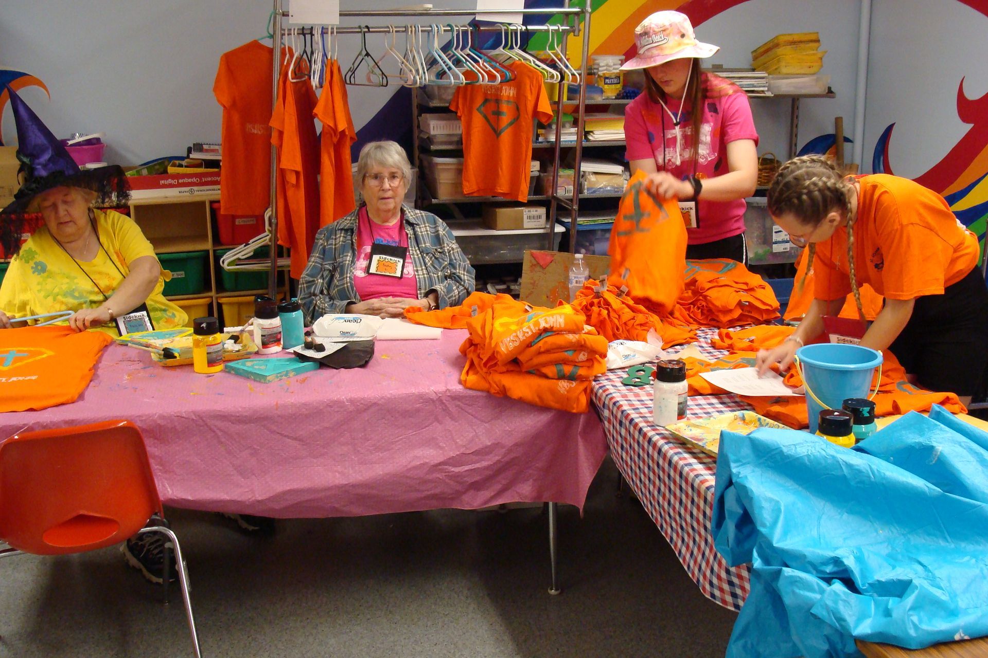 People folding orange shirts at a table in a workshop with colorful fabric and supplies