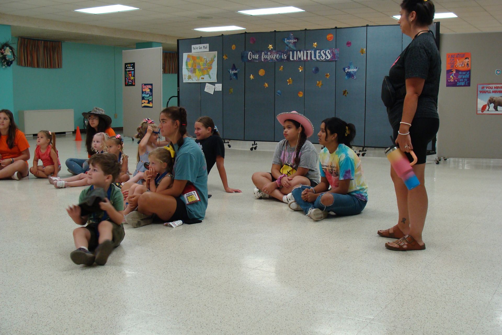 Children sitting on the floor in a classroom while a teacher stands nearby