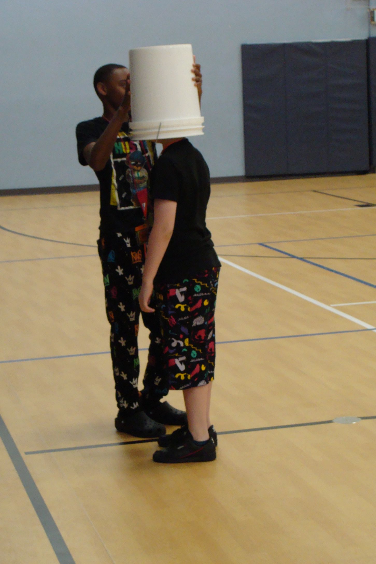 Two people standing face to face in a gym, one wearing a white bucket on their head