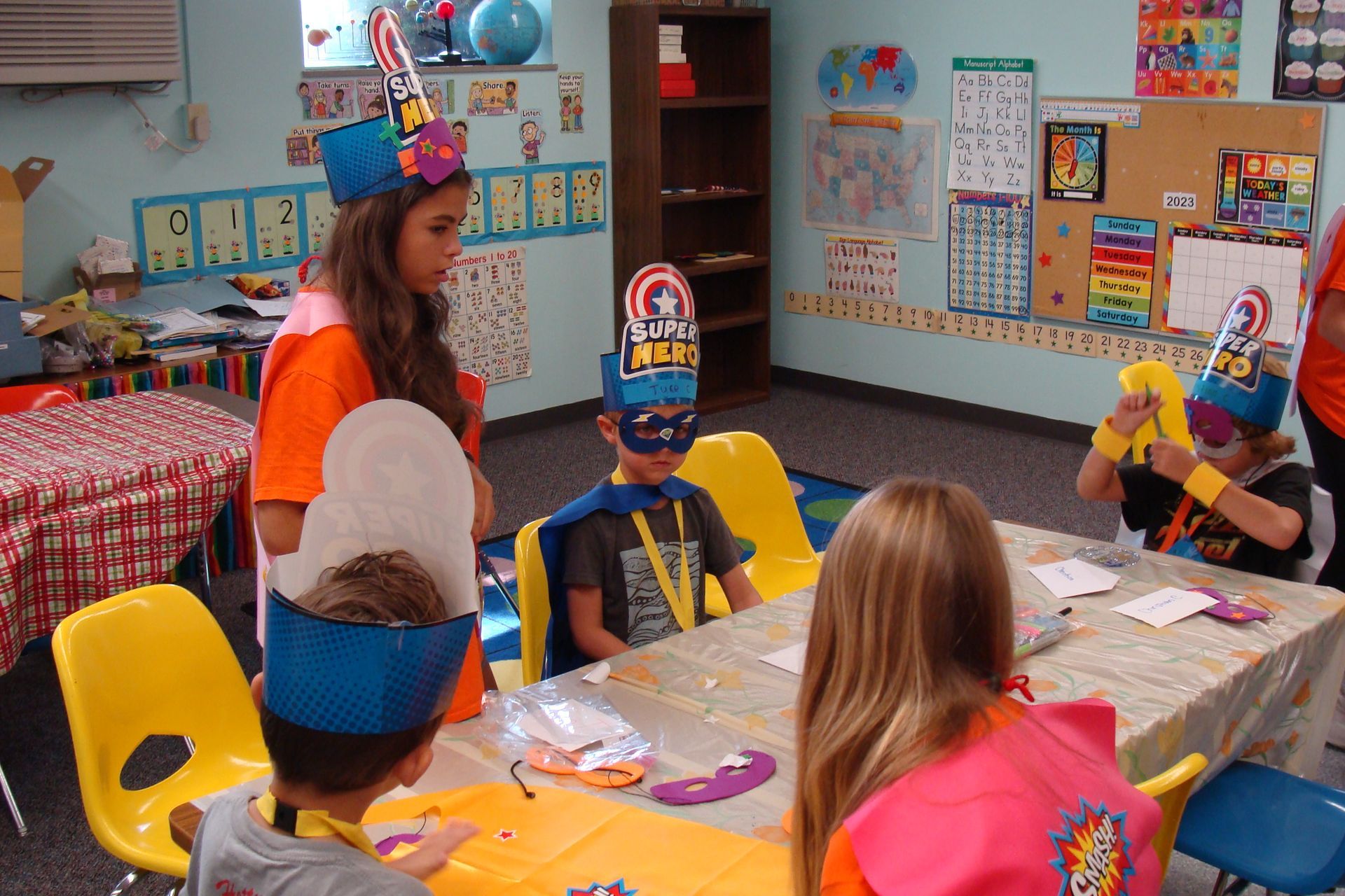 Children in party hats sit at a classroom table while a woman in an orange shirt stands nearby.
