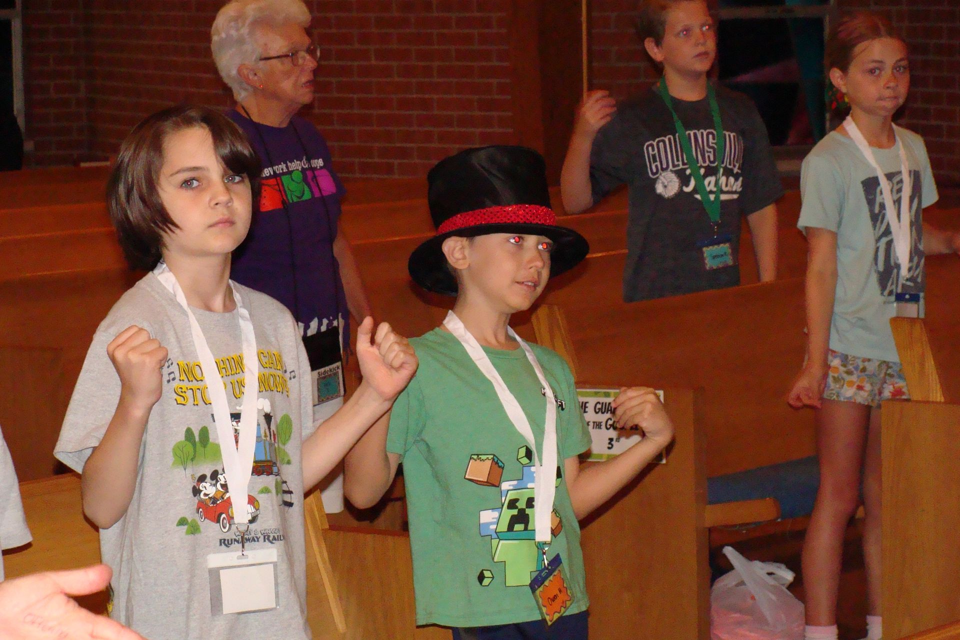 Children standing in a wooden hall, some wearing lanyards and playful hats, as if in a group activity.
