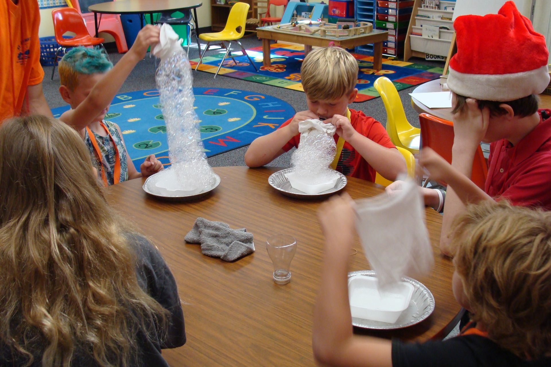 Children do science at a classroom table, watching white foam erupt from cups.