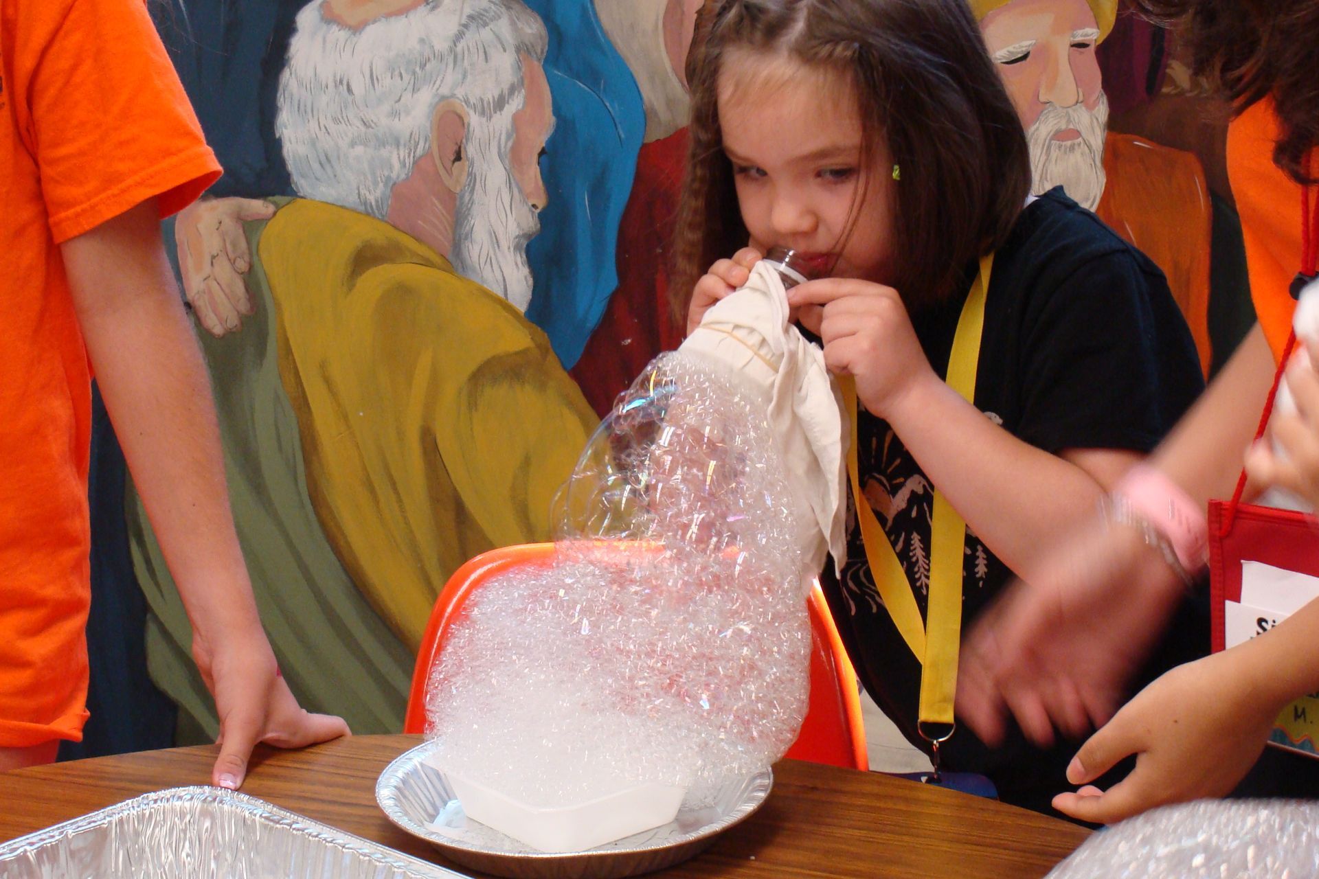 Child in black shirt blowing into a bag of foam beads at a table, with adults nearby and a mural behind.