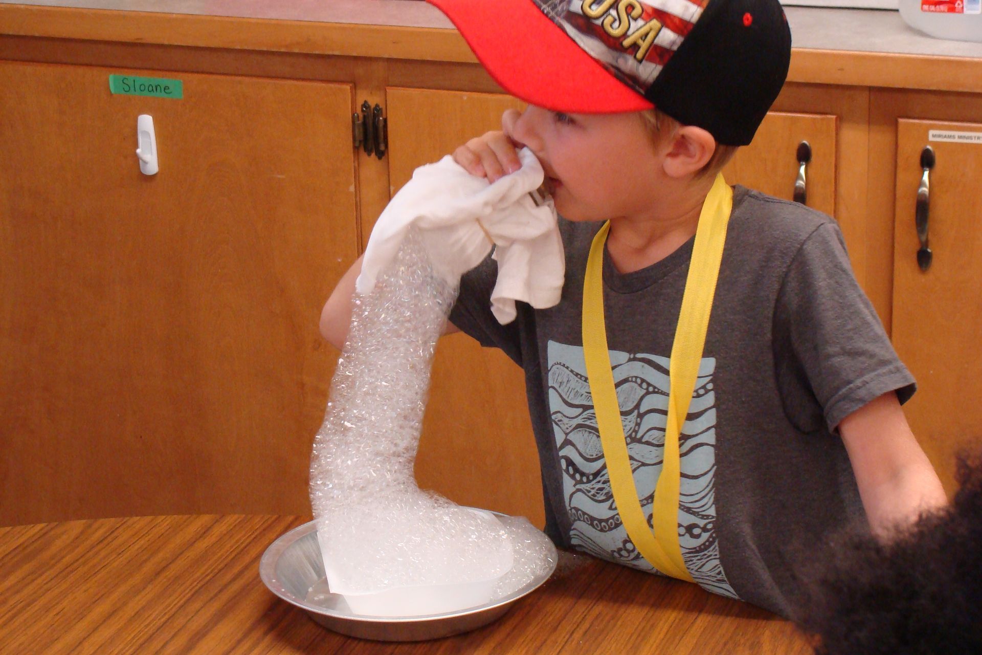 Child in a red cap and yellow suspenders blowing a foamy white experiment in a bowl on a table