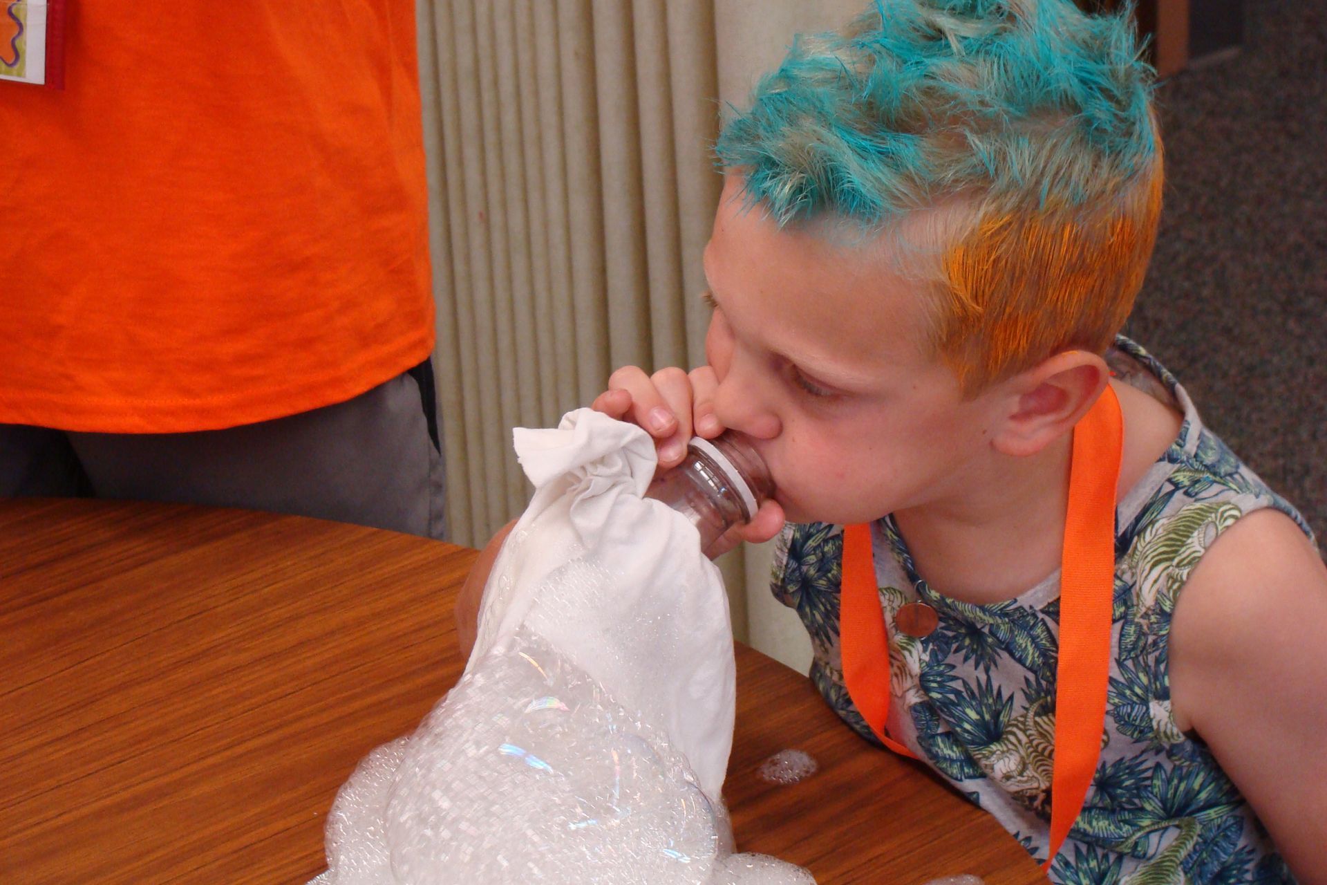 Child with blue and orange hair drinking from a bottle at a table with a bag of ice