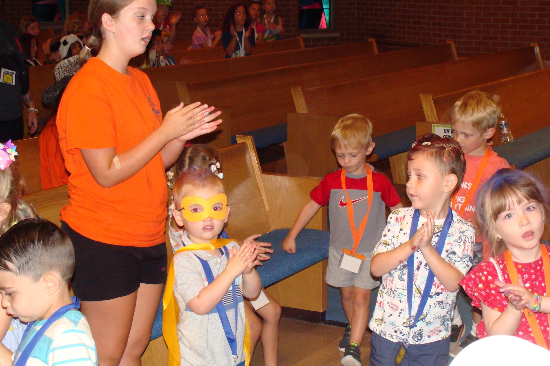 Teacher in orange shirt leading children in a classroom activity on a stage-like floor