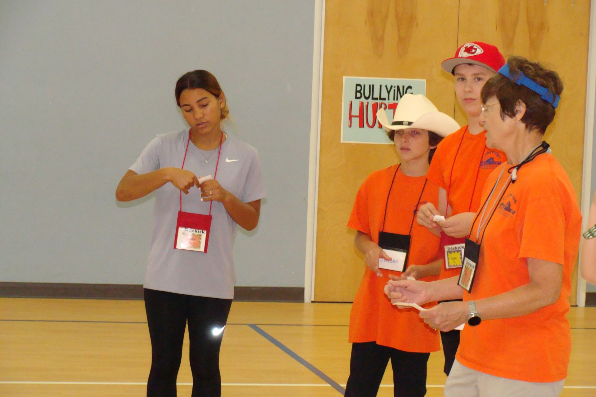 People in orange shirts and caps standing indoors during a group activity, with one person reading from notes.