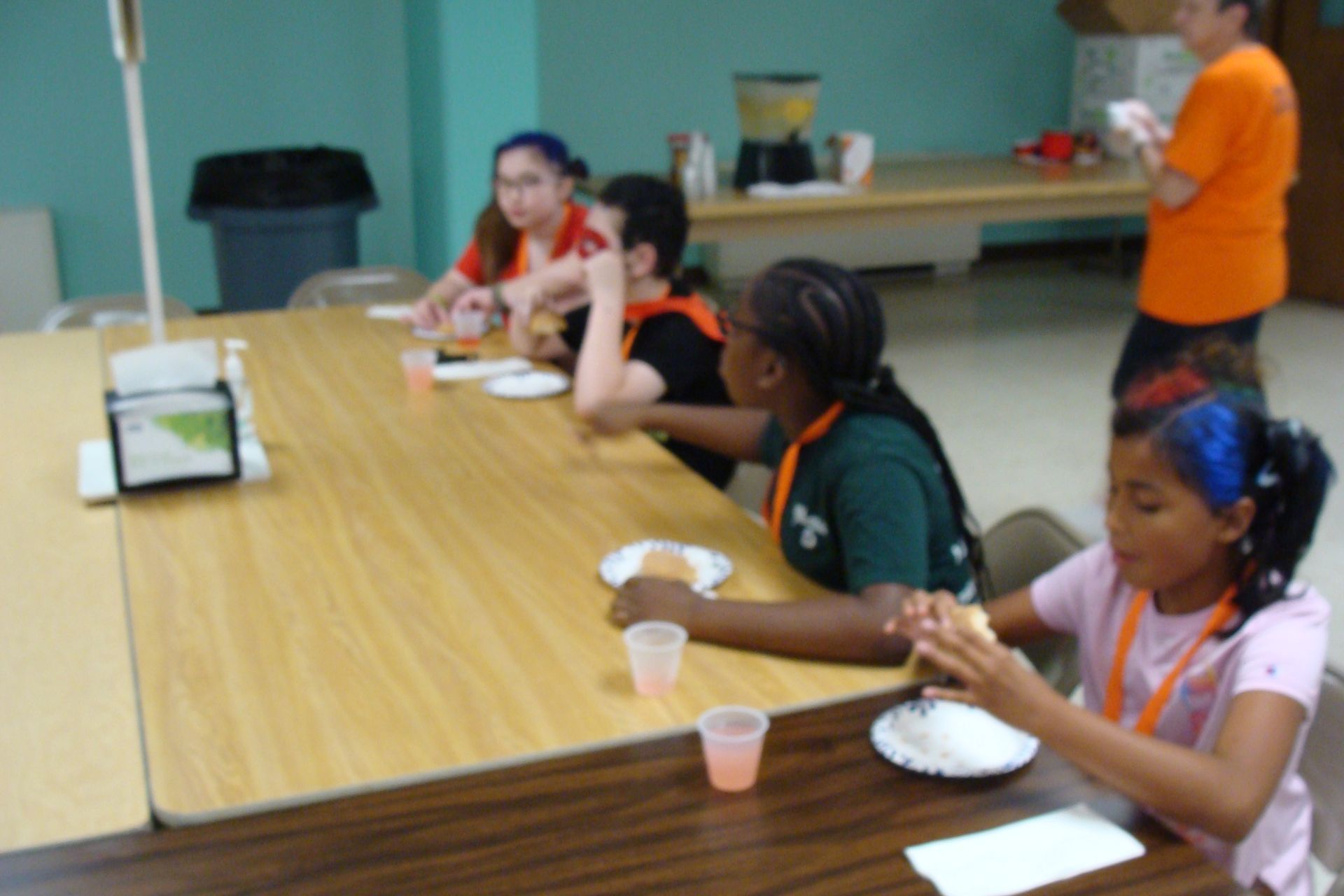 People seated at a cafeteria table eating plates of food, with an adult standing nearby.